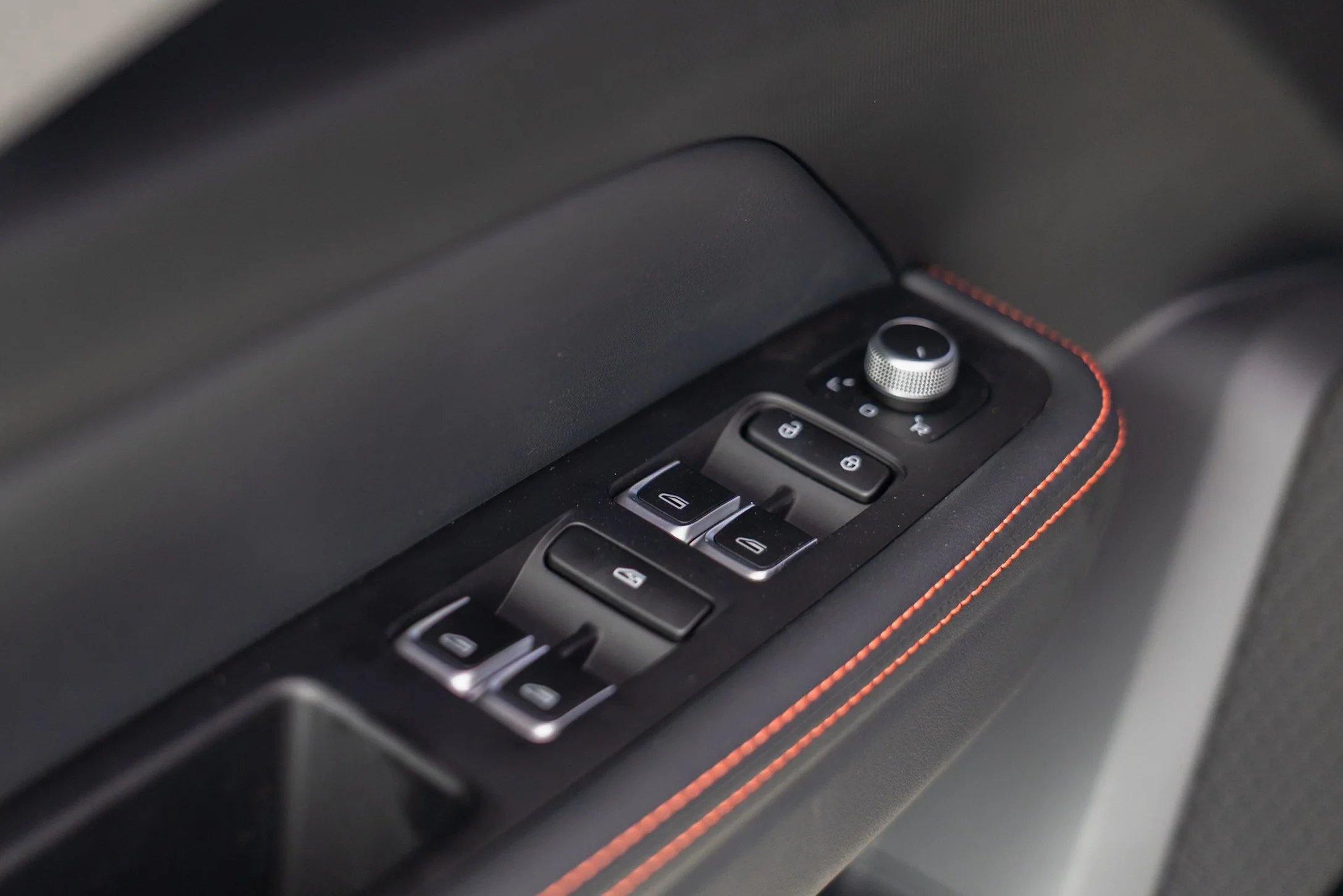 Close-up of a car's interior door panel, showing window control buttons, a rotary dial, and red stitching on the leather surface.