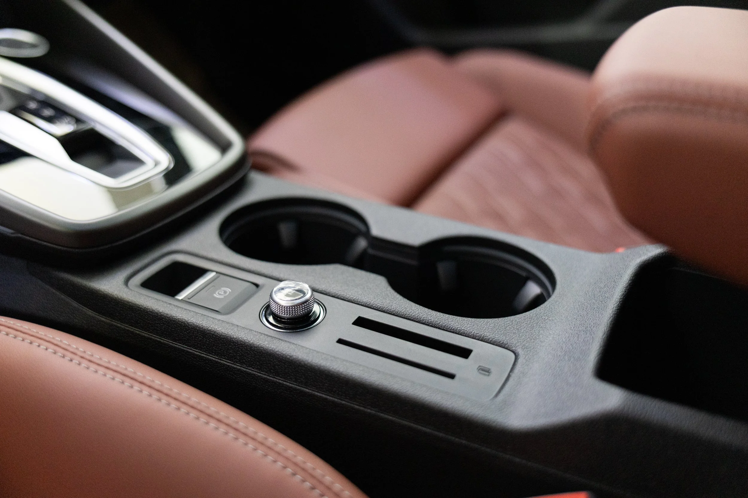 Close-up view of a car's center console with gear shift, electronic parking brake button, and cup holders, surrounded by brown leather seating.