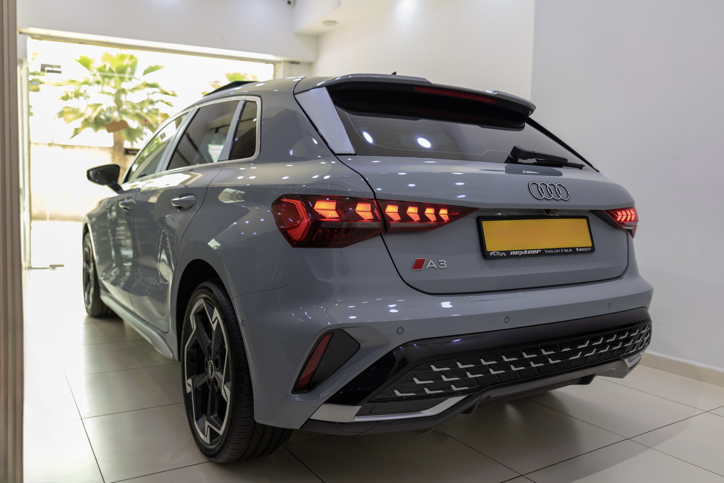 A silver Audi A3 hatchback parked indoors near a large window with a blurred green plant outside. The car features LED taillights, a rear spoiler, and a black grille, with a dealership or service sticker on the rear bumper.