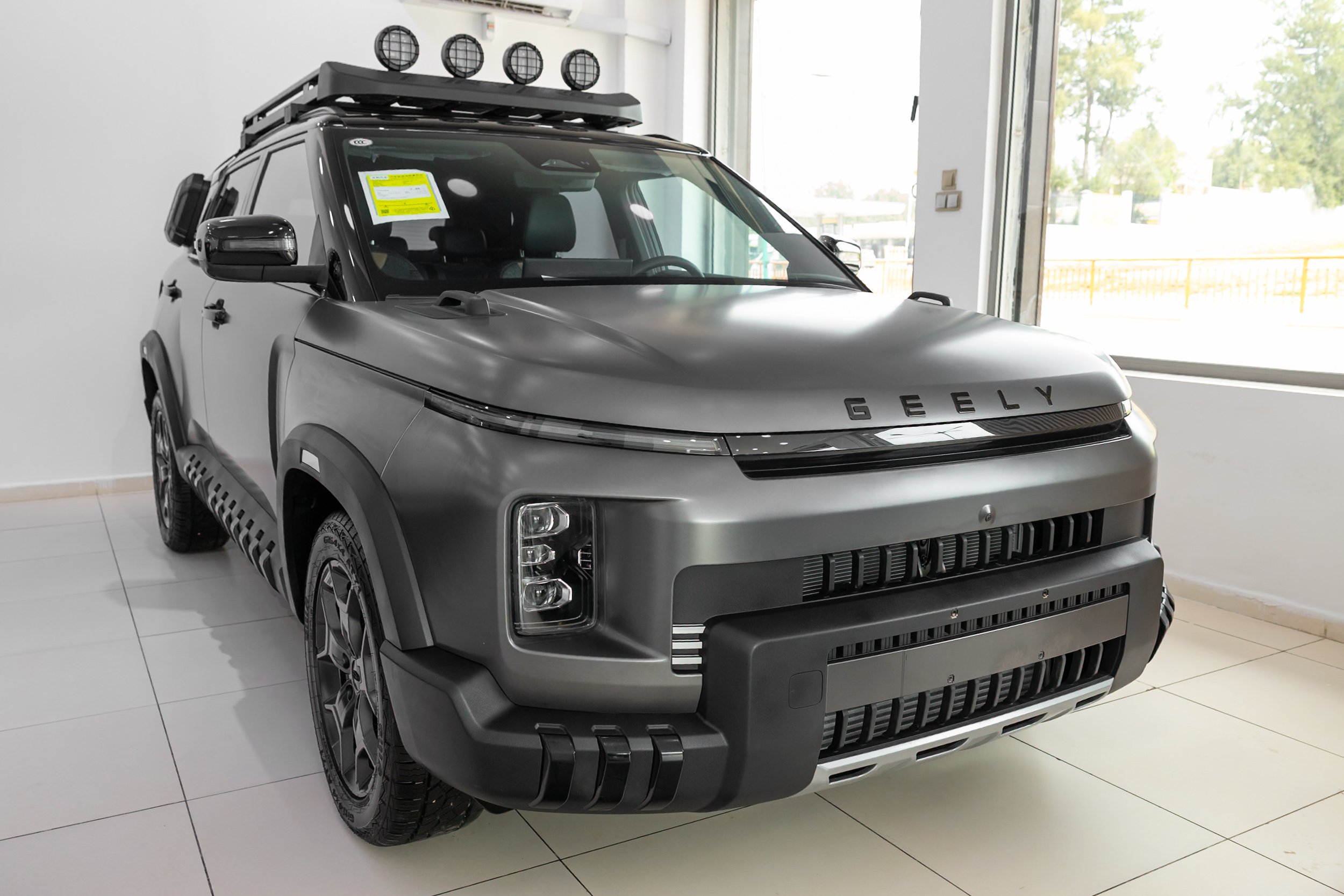 A modern, gray Geely SUV displayed indoors next to a large window, with a roof rack and five spotlights on top.