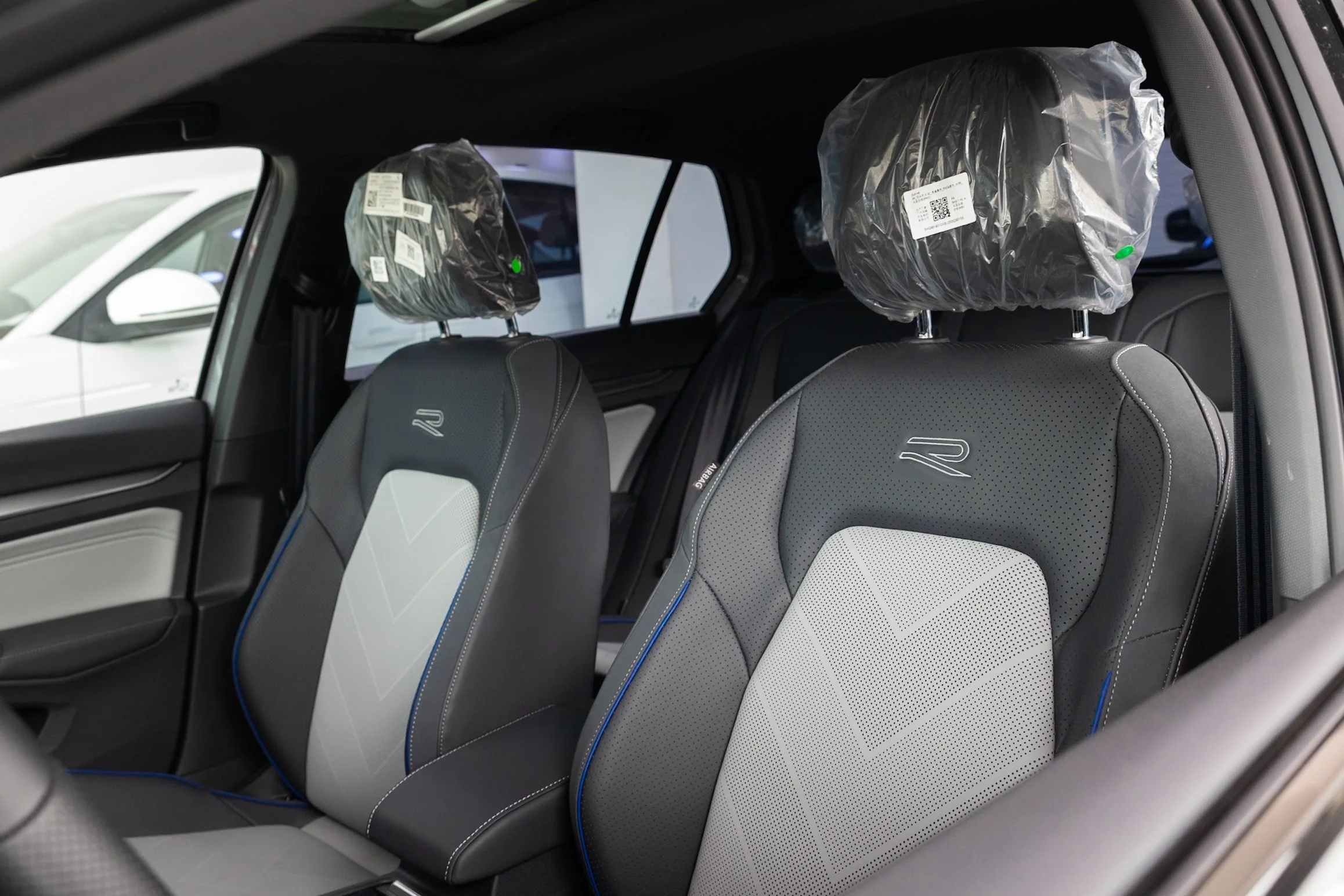 The image shows the interior of a car, focusing on the back seats. The headrests are covered with plastic, indicating the vehicle might be new. The seats are gray with a patterned design and blue piping. There are windows visible, with one showing a 