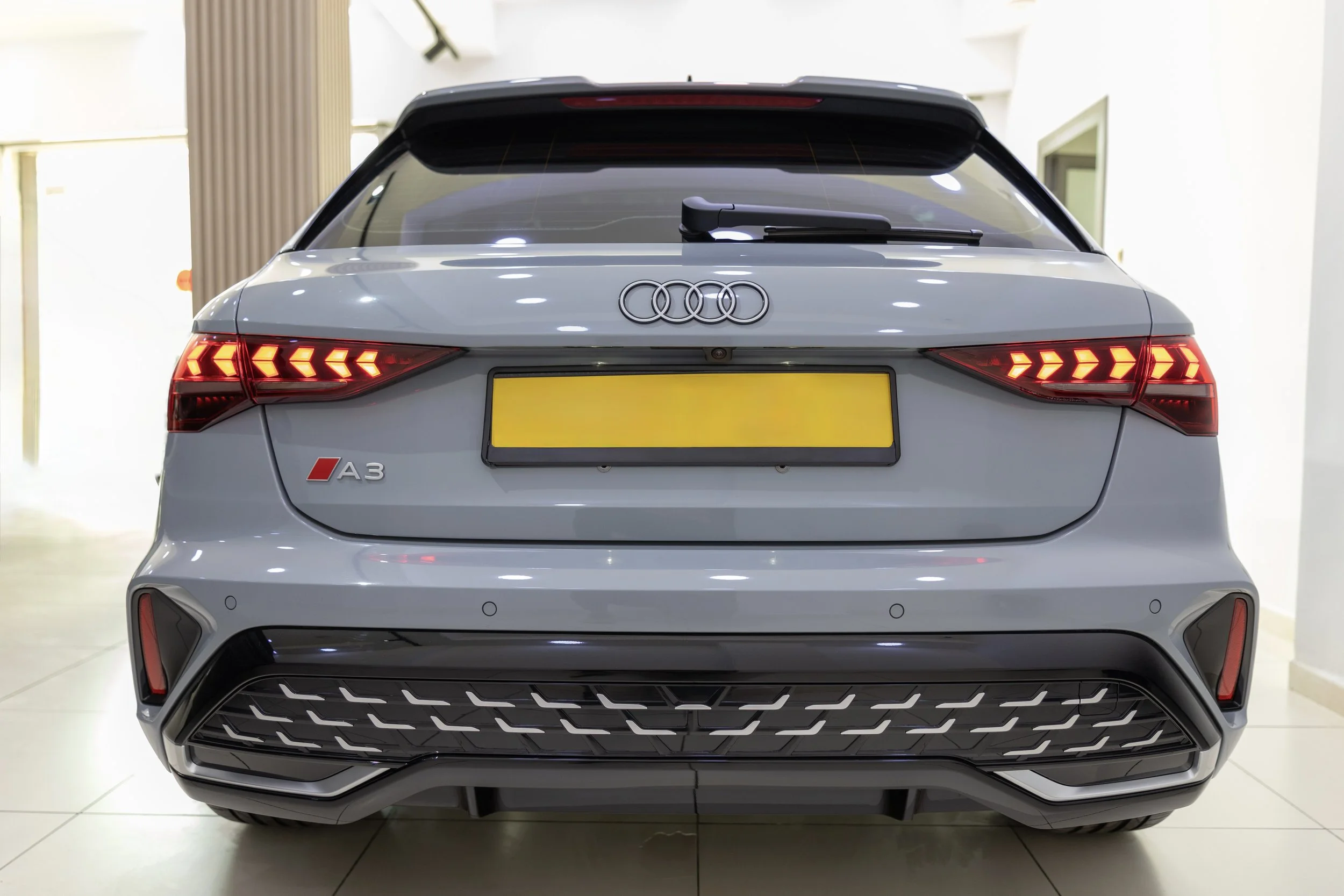 The rear view of a white Audi A3 hatchback car inside a building, showing illuminated taillights, a black grille at the bottom, a rear windshield wiper, and the Audi logo on the back.