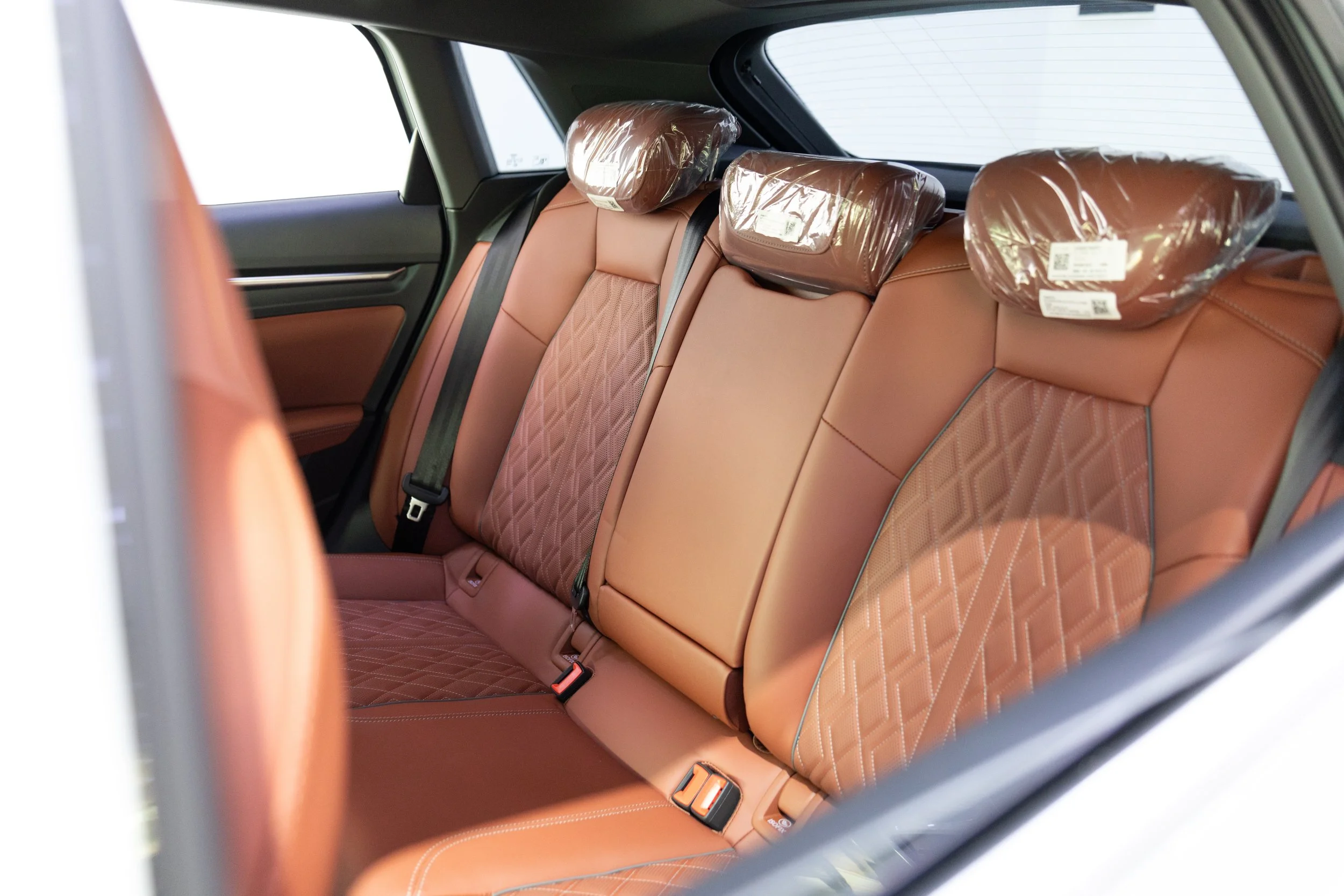 The back seat of a car showing tan leather seats and headrests covered in plastic.