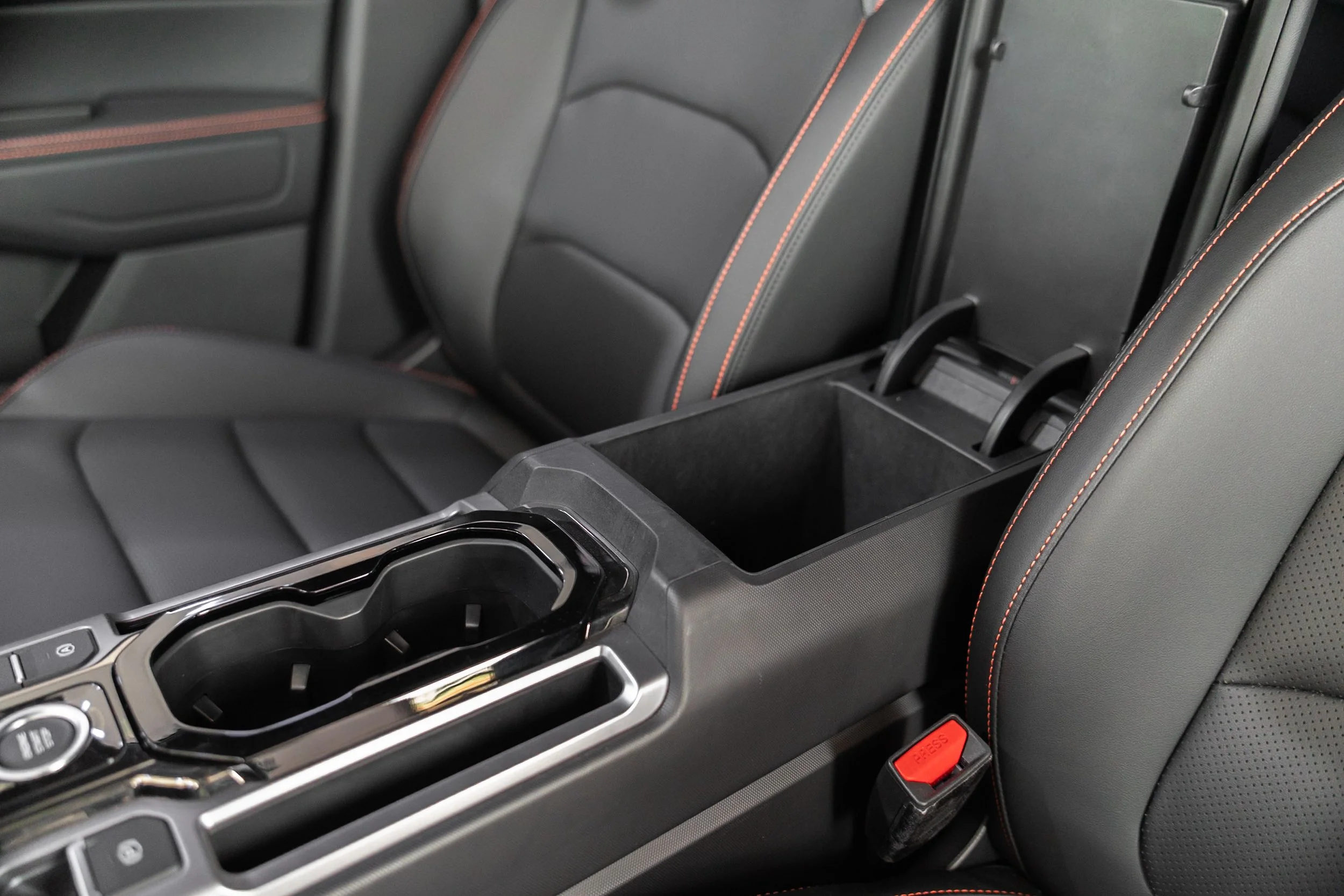 Close-up of the center console and back seat of a modern car interior, with black leather seats accented by red stitching, a cup holder, and a red 'press' button.