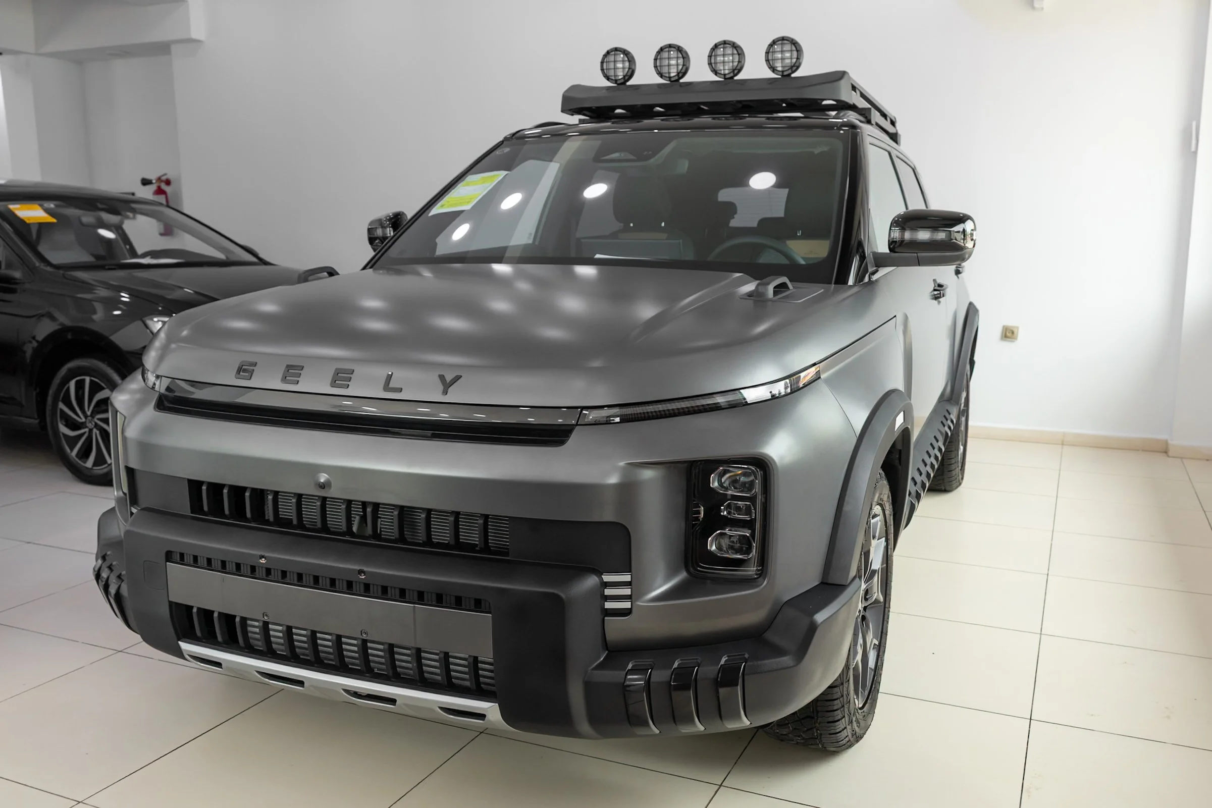 A silver Geely vehicle with off-road features on display in a showroom, accompanied by a black car, white walls, and tiled floor.