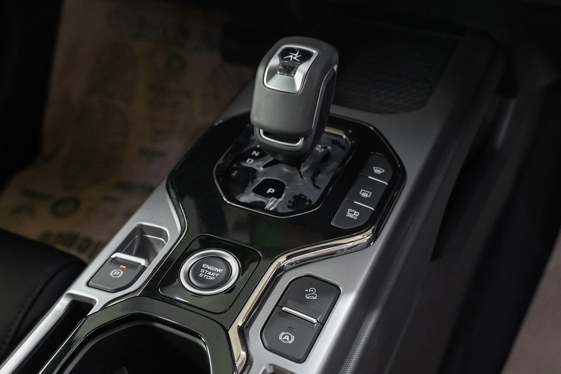Close-up of a modern car's center console with an electronic gear shift, engine start/stop button, parking brake, and other control buttons.
