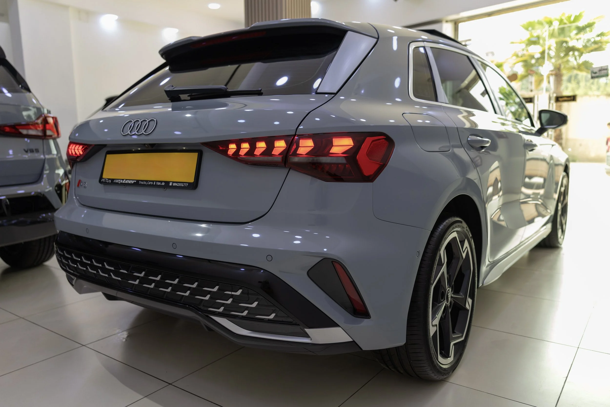 Rear view of a silver Audi hatchback in a showroom with two other cars visible, showcasing distinctive taillights and modern design.