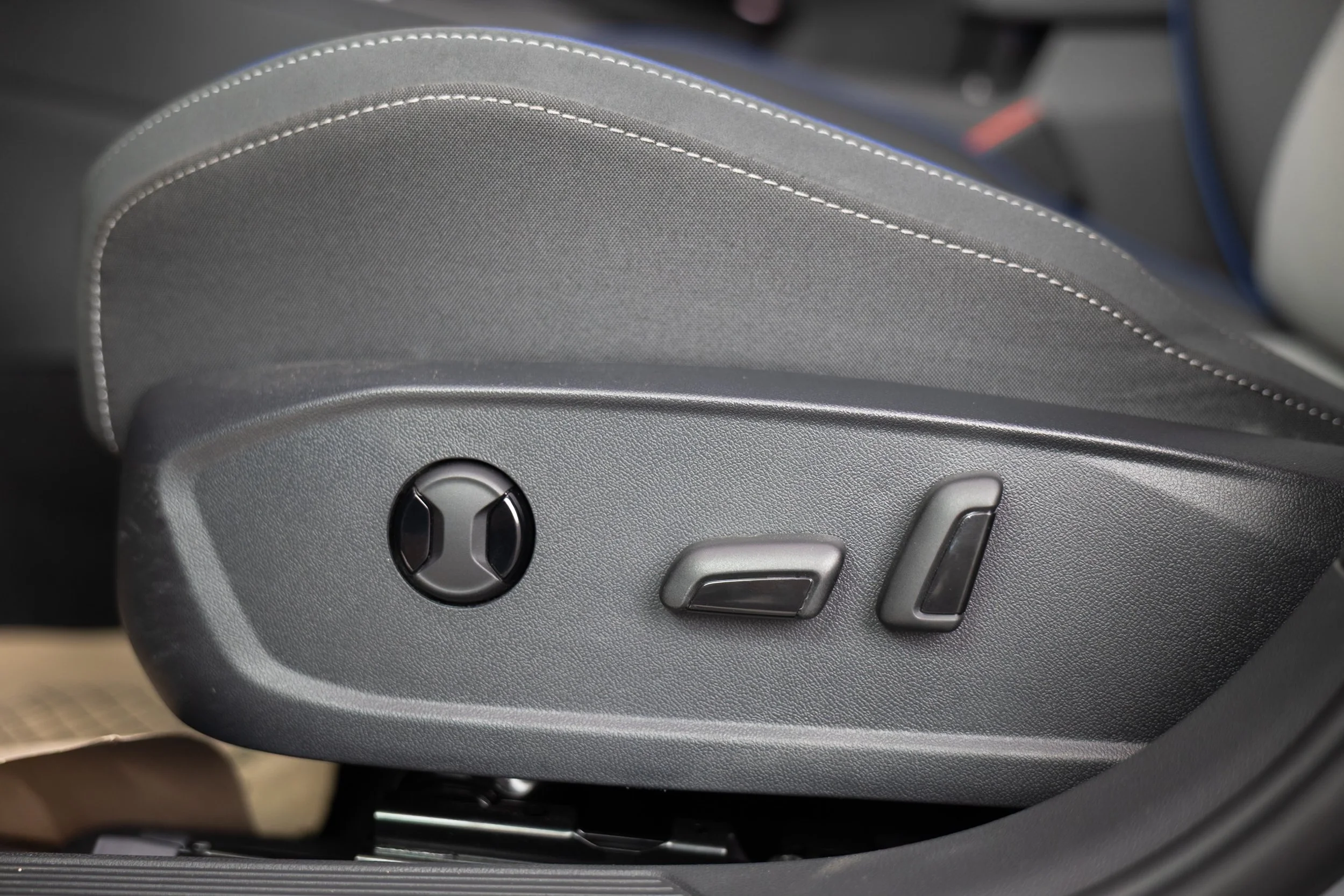 Close-up of the side of a car seat showing adjustment controls, including a circular knob and two levers on a textured plastic panel.