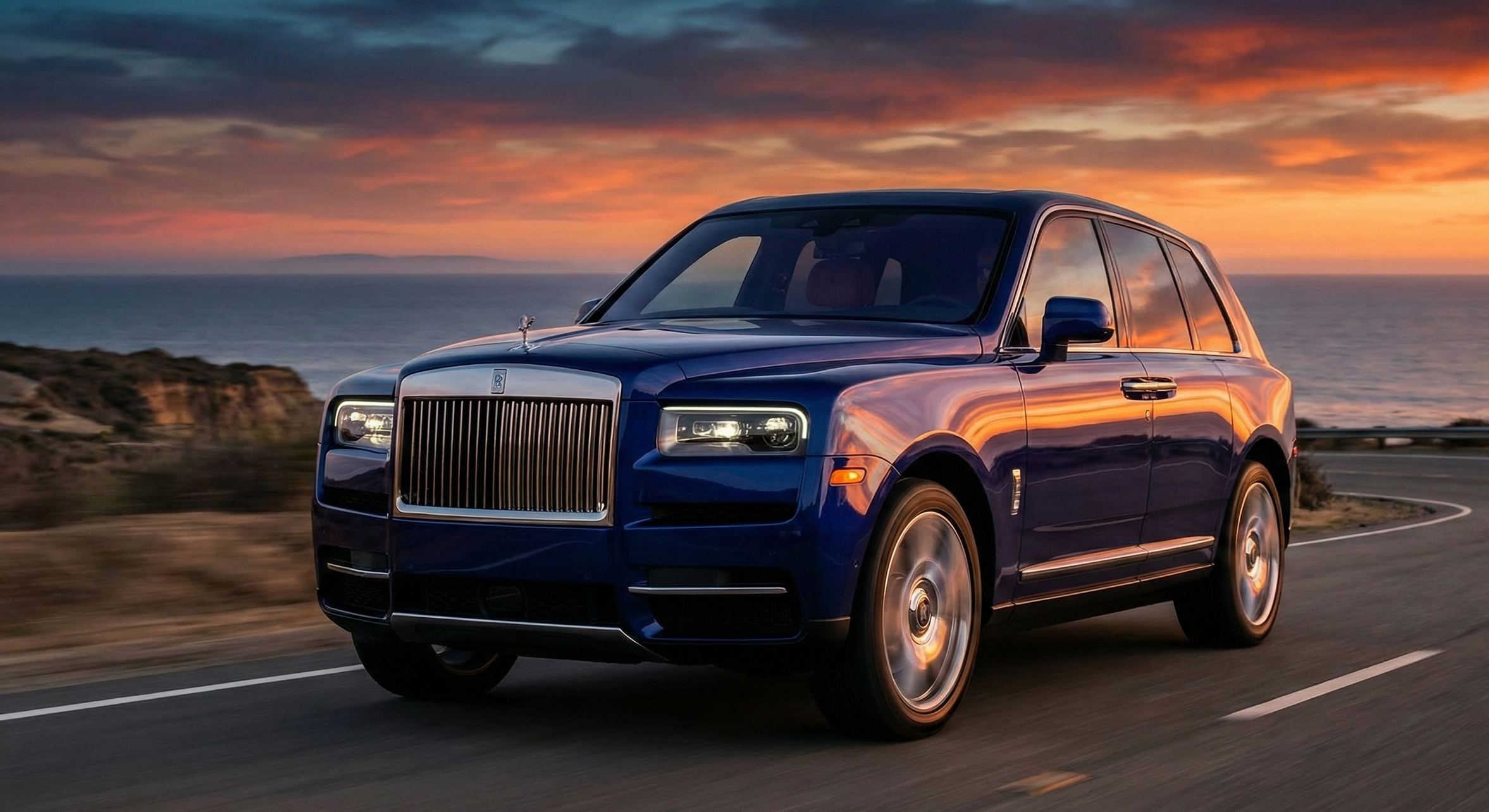 Une Rolls Royce Cullinan bleue élégante roulant le long d'une route au coucher du soleil avec la mer en arrière-plan.