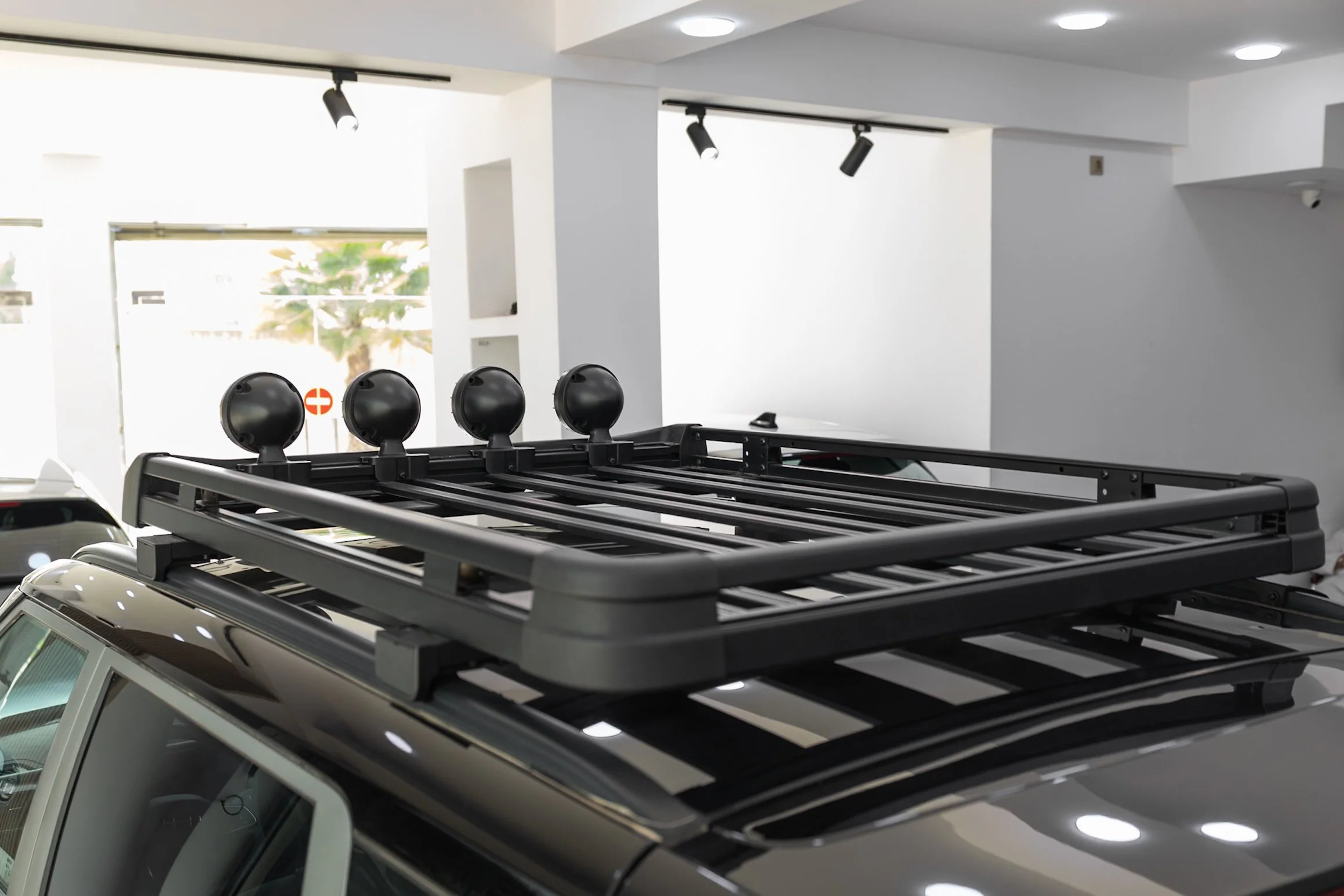 The image shows the roofs of multiple parked cars within a covered parking garage. There is a roof rack installed on top of the first car visible in the foreground, featuring four round lights attached.