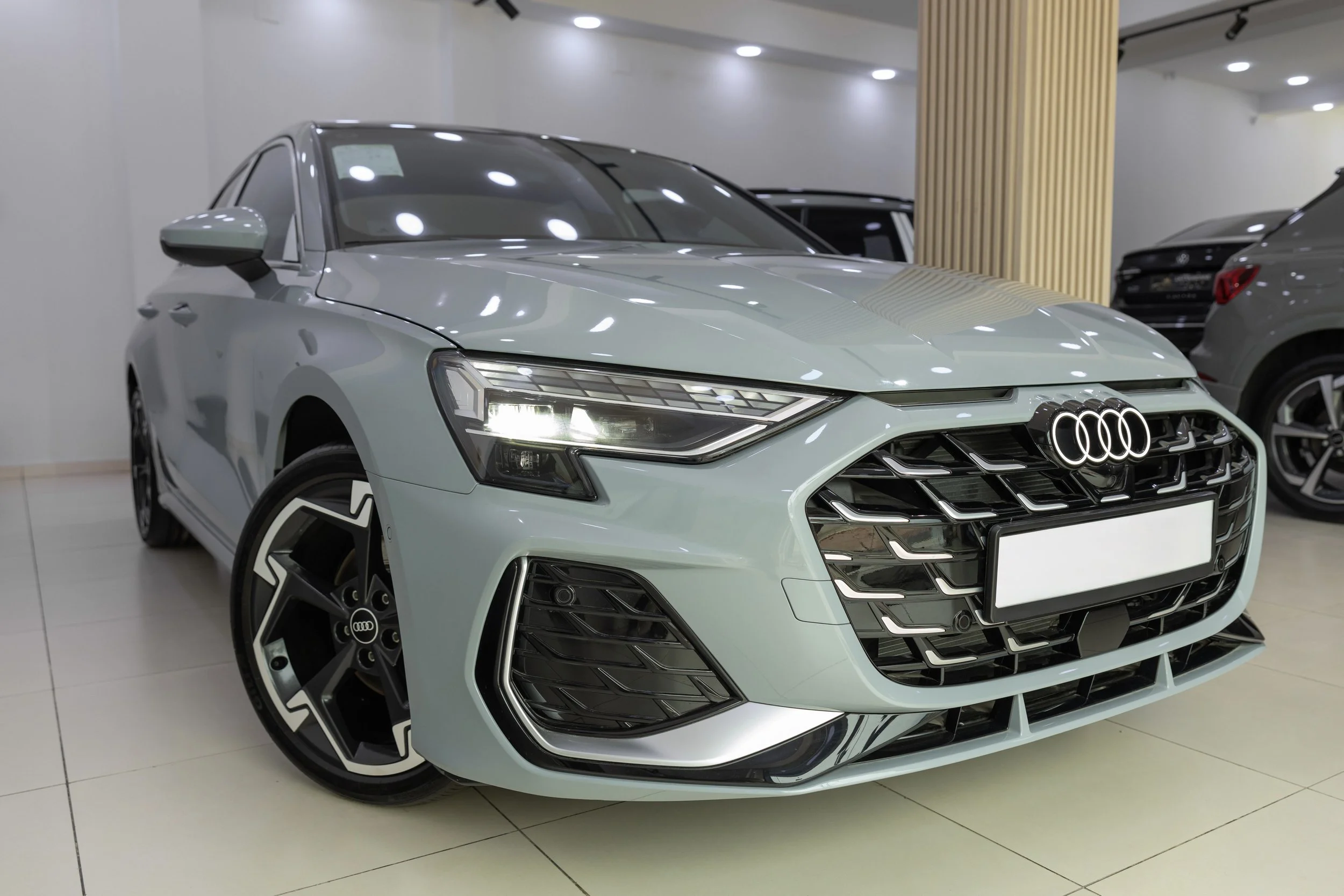 A silver Audi sedan on display in a showroom, with black and silver wheel rims, headlights on, and a modern interior with white walls and ceiling lights.