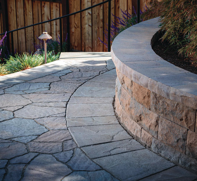 Curved stone patio with a low stone retaining wall, outdoor garden lighting, and plants along a wooden fence.