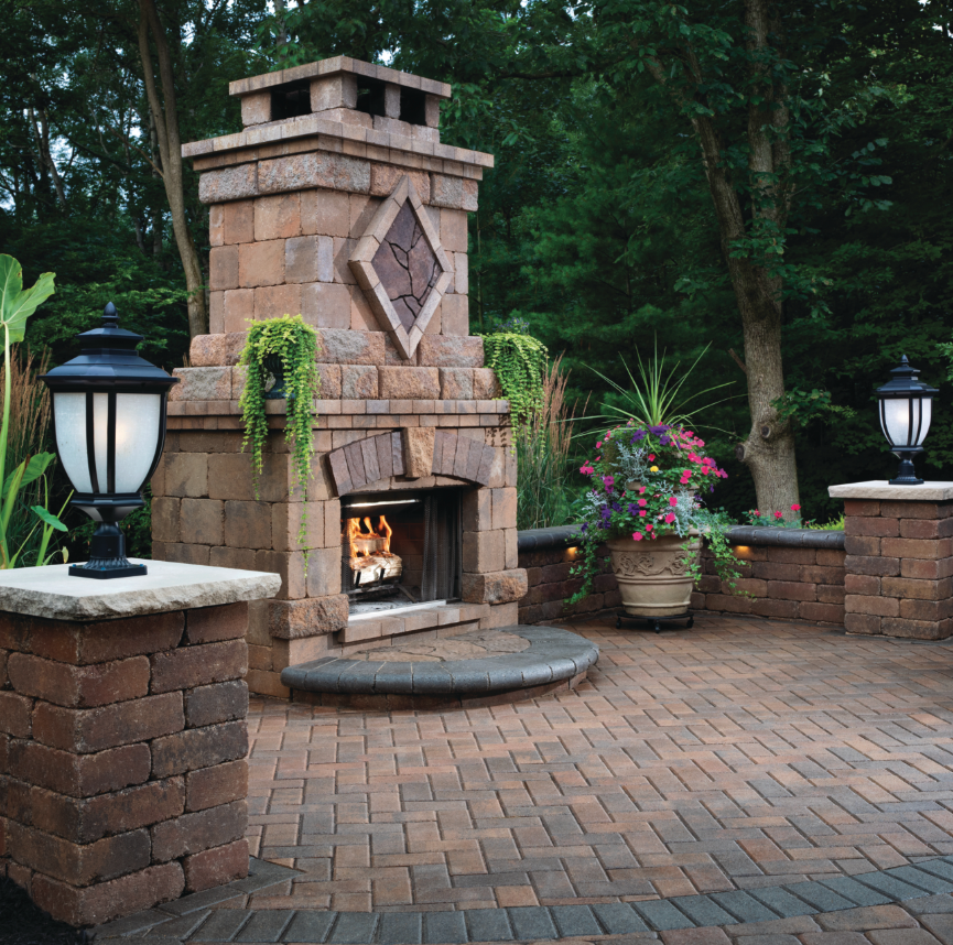 Outdoor patio with a stone fireplace, brick flooring, and lanterns on brick pedestals. Potted plants with green foliage and colorful flowers decorate the area, surrounded by trees.