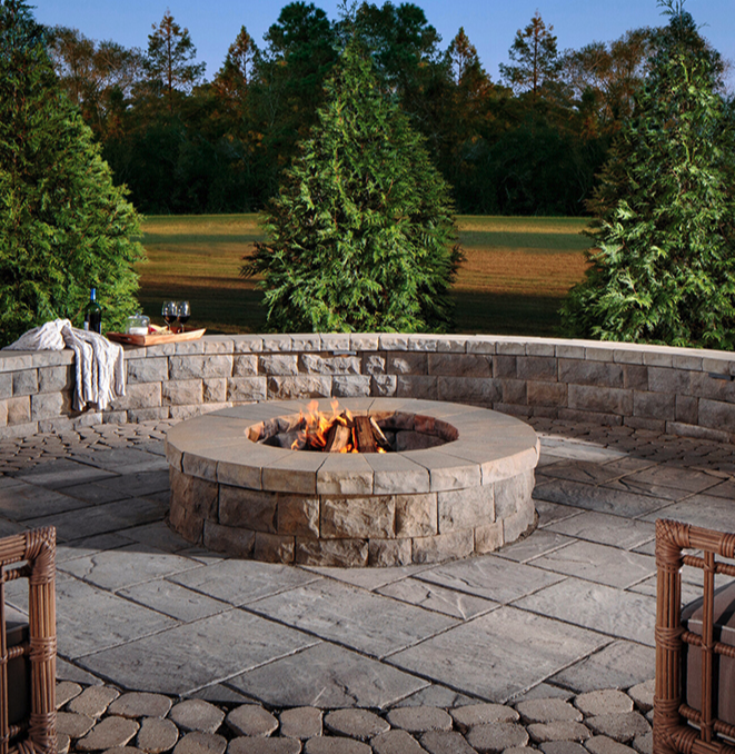 Outdoor patio with a circular stone fire pit, surrounded by stone and brick flooring, in a backyard with tall evergreen trees and a lawn.