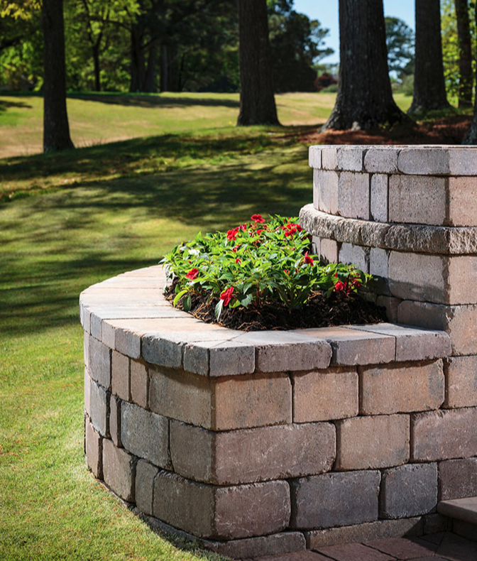 A brick planter with red and green flowering plants on a grassy lawn with trees in the background.