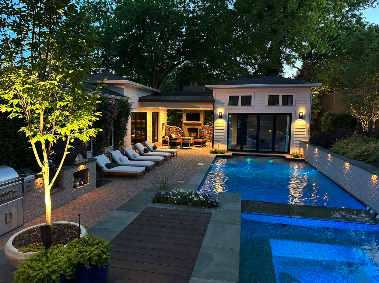 Upscale backyard with a swimming pool, lounge chairs, outdoor fireplace area, trees, and a small patio house during evening.