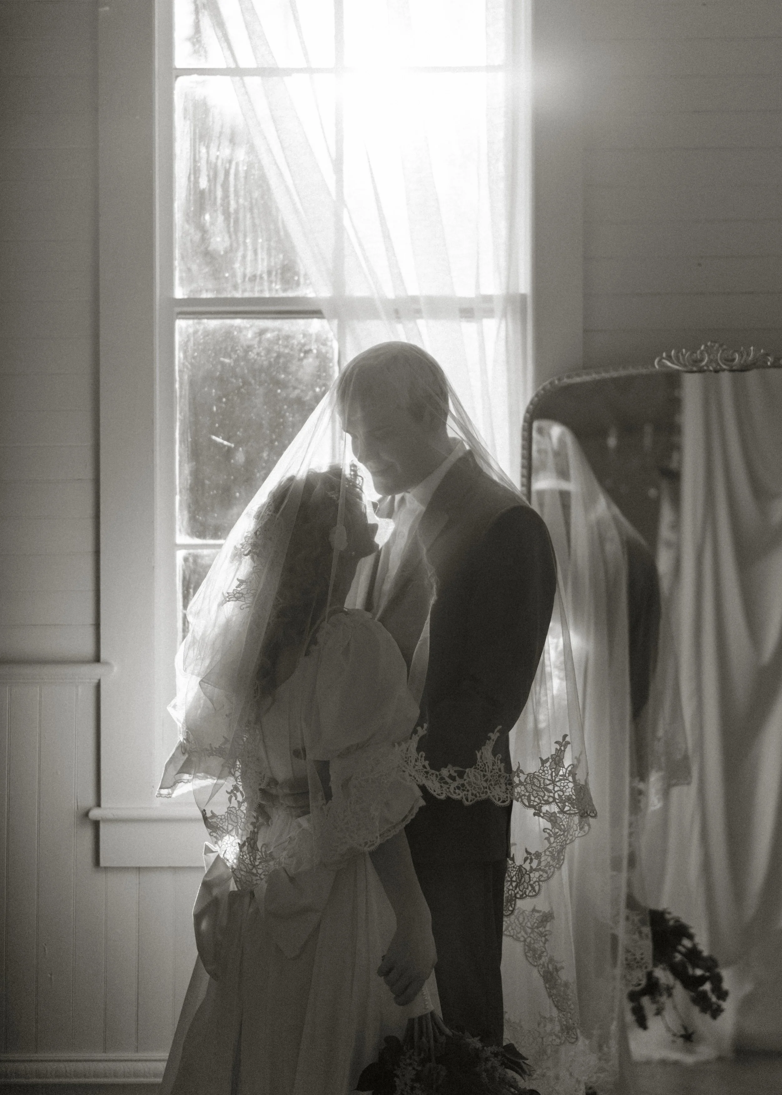 A black-and-white photo of a bride and groom inside a room, partially illuminated by sunlight coming through a window. The bride is holding a bouquet and wearing a lace wedding dress with a veil, while the groom is in a suit, with their foreheads and