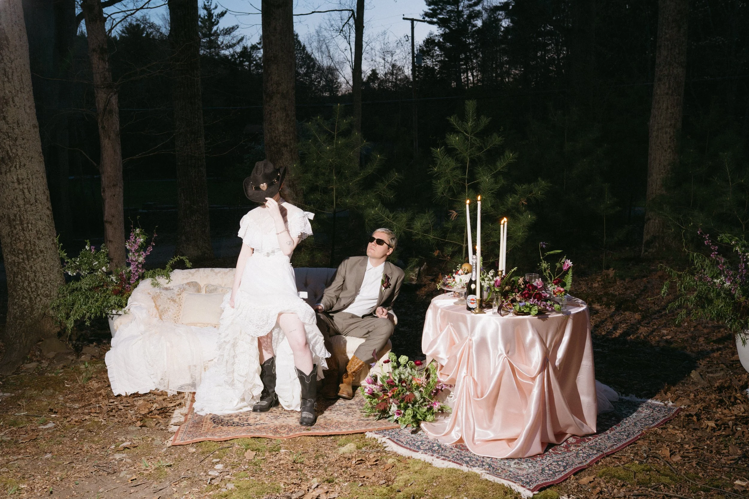 A couple dressed in vintage attire sitting outdoors at night, surrounded by trees, with a decorated table and floral arrangements, candles, and drinks.