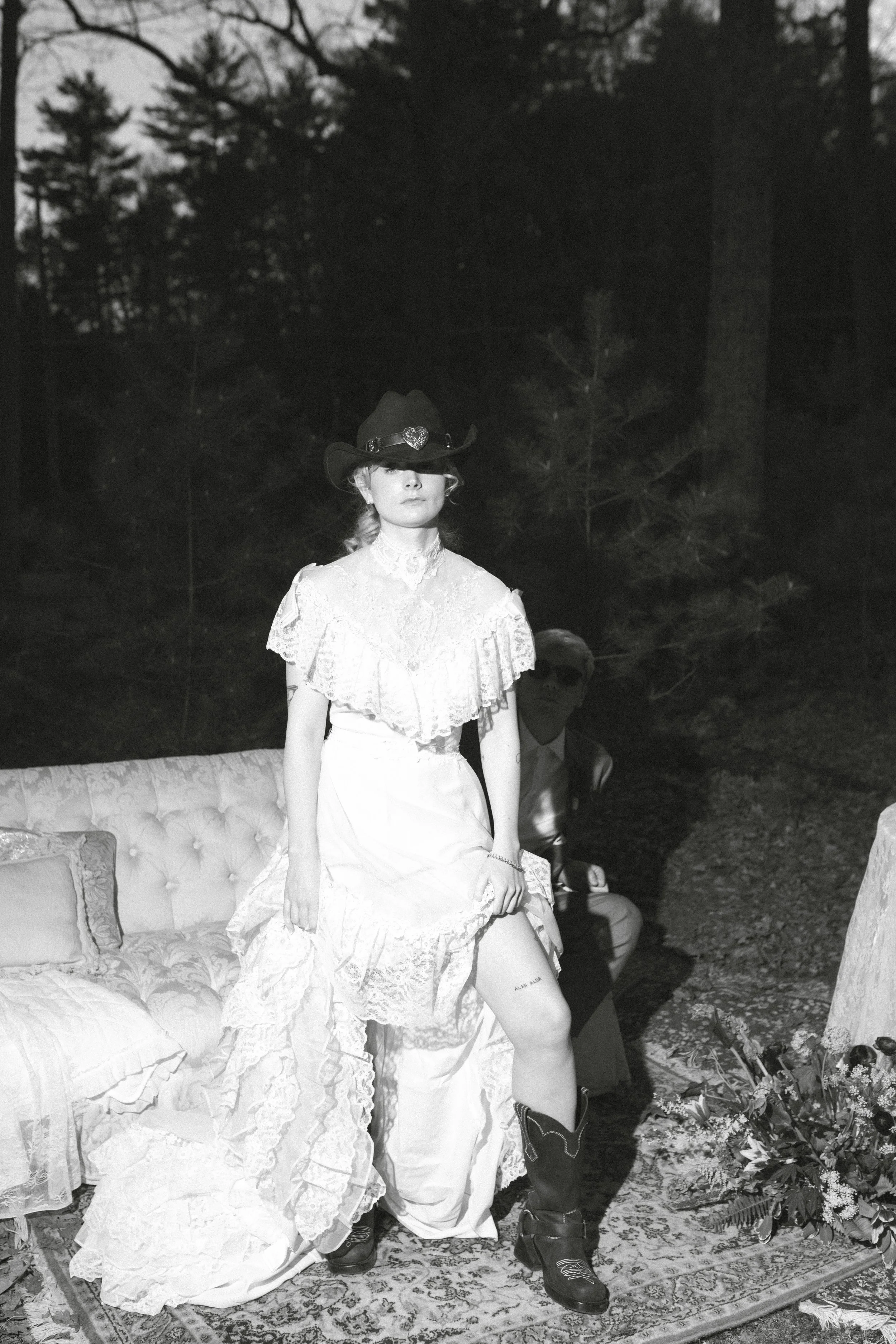 A woman wearing a lace dress, cowboy hat, and boots standing outdoors near a sofa with floral pillows and a bouquet of flowers on the ground, with a man sitting behind her in a forest setting.