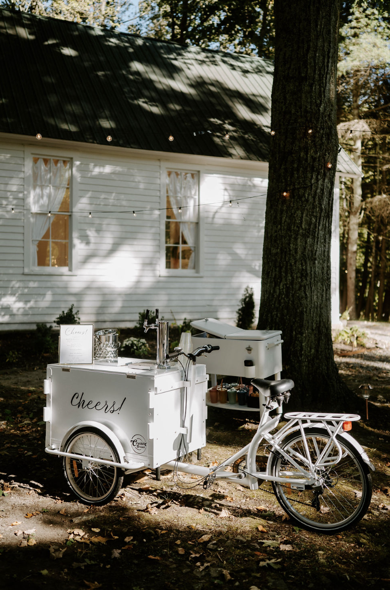 A white tricycle with a drink cart attached, labeled 'Cheers!', parked outdoors in a shaded area with a house and trees in the background.
