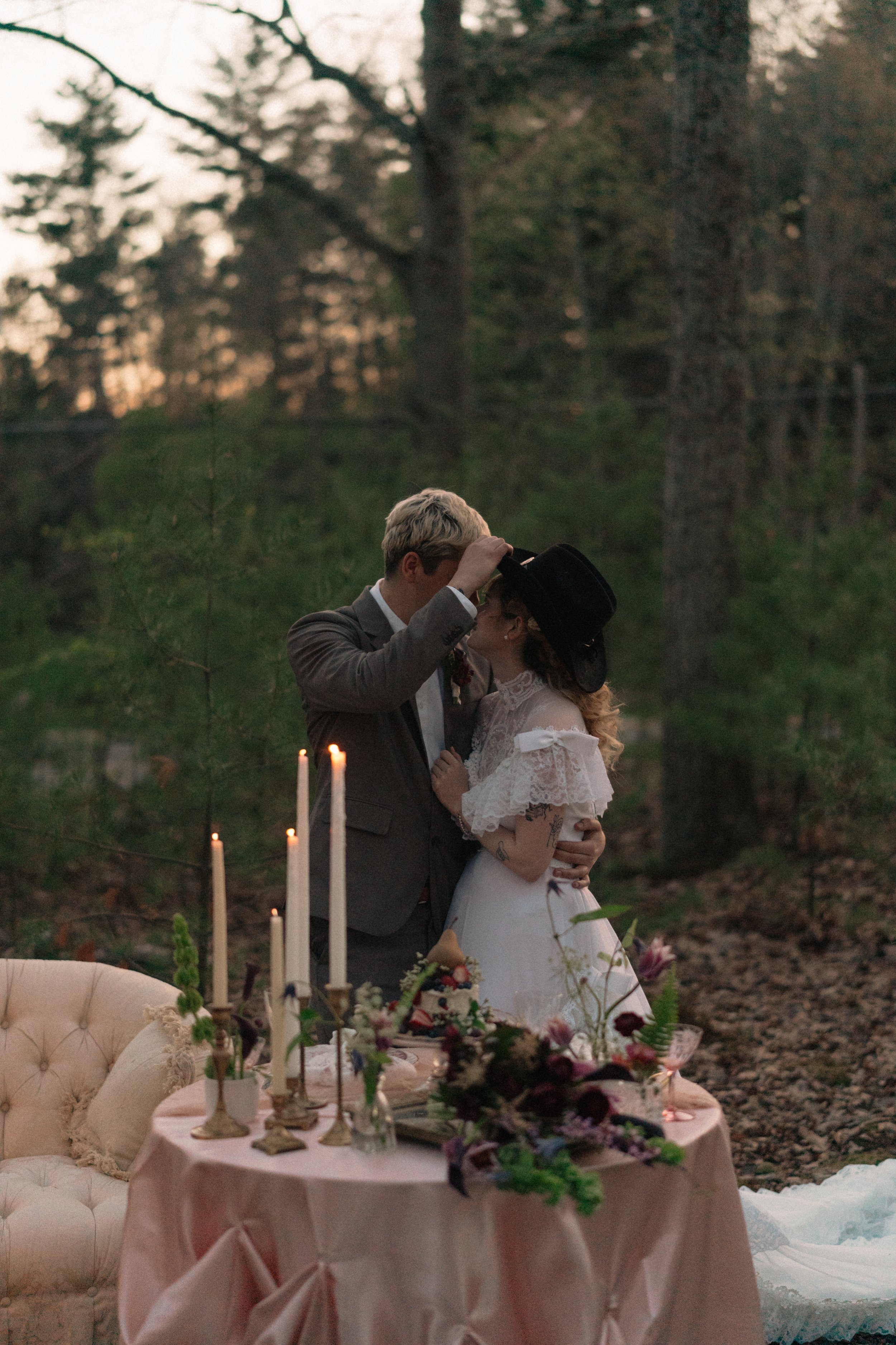 A couple dancing romantically outdoors at a wedding reception around sunset, with a table adorned with flowers, candles, and a cake in the foreground.
