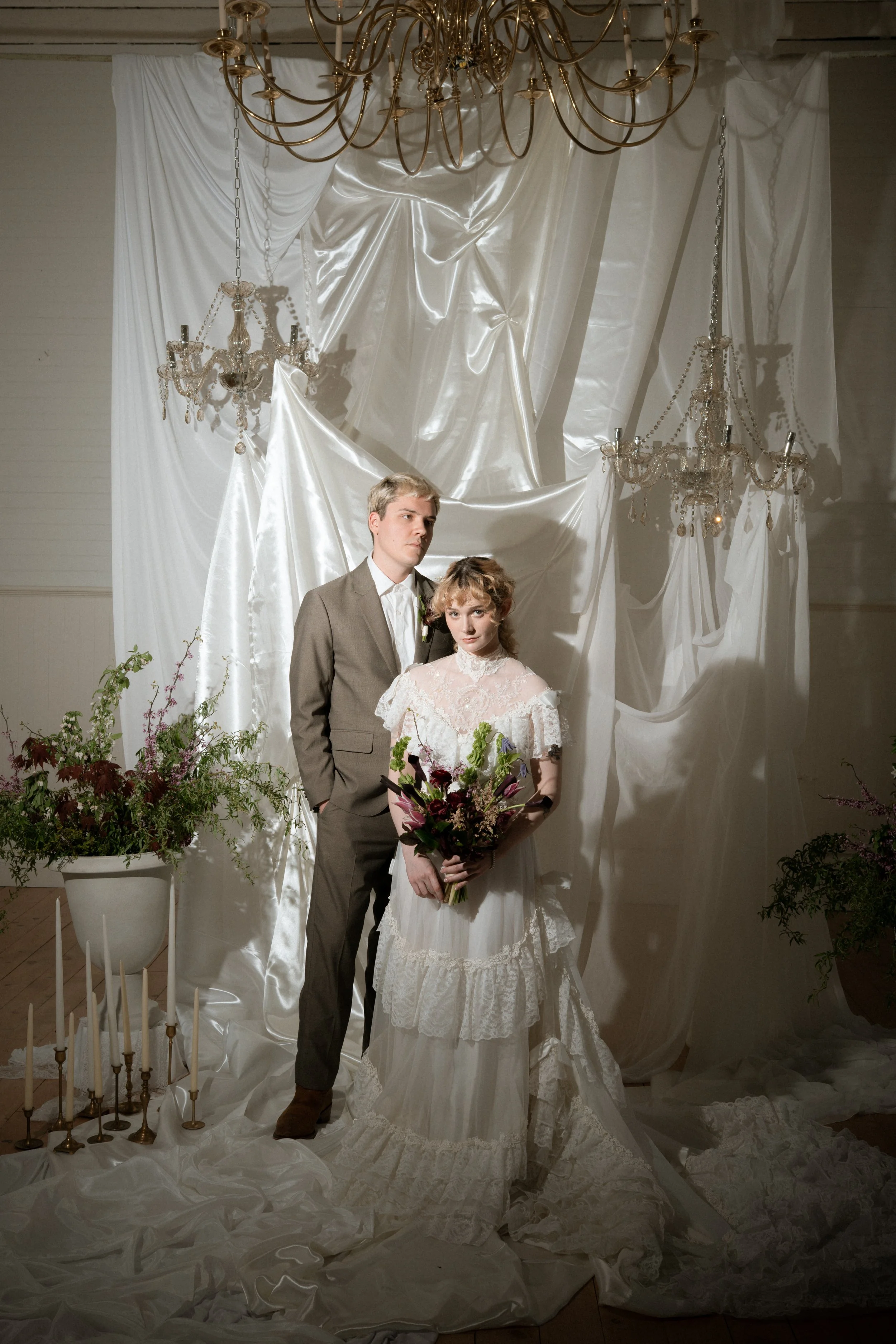 A bride and groom standing together at a wedding portrait, with the bride holding a bouquet and the bride wearing a lace wedding dress, in front of a backdrop with white fabric curtains, chandeliers, and floral decorations.