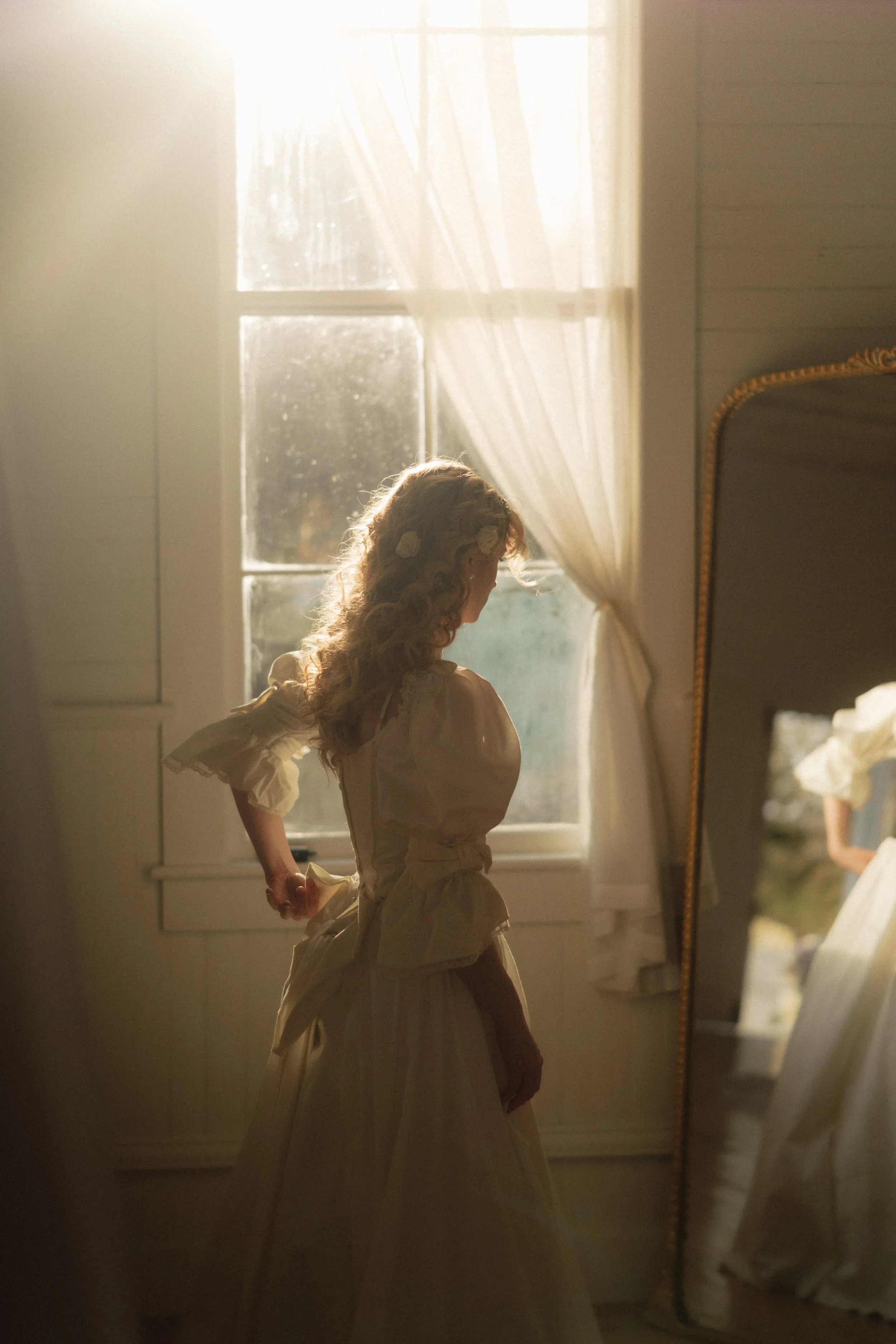A woman in a vintage-style white dress and curly hair stands in front of a window with sheer curtains, backlit by sunlight, with her reflection visible in a large standing mirror.