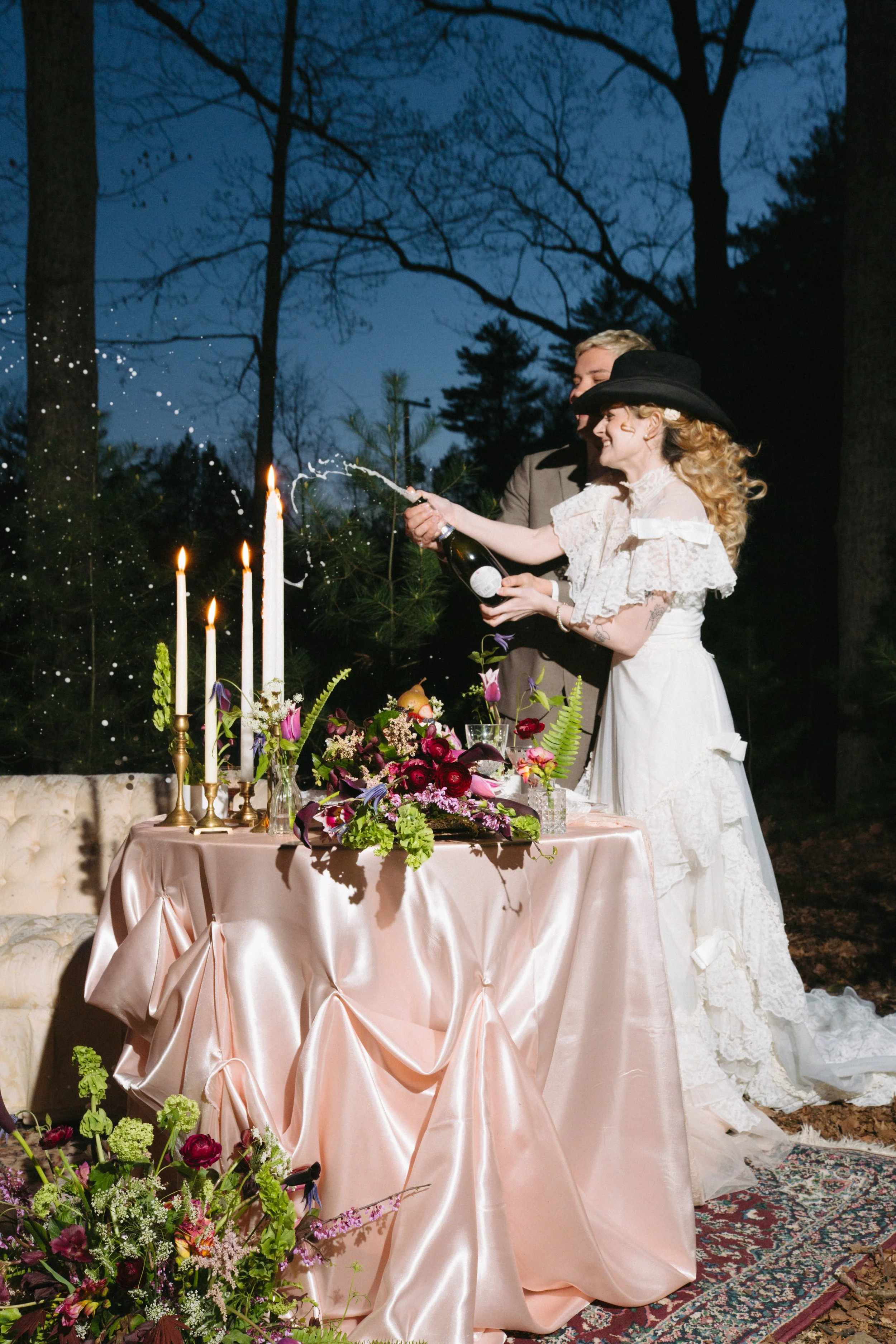 A woman and man celebrate outdoor wedding with champagne, surrounded by floral arrangements and candles on a pink satin-draped table at dusk.