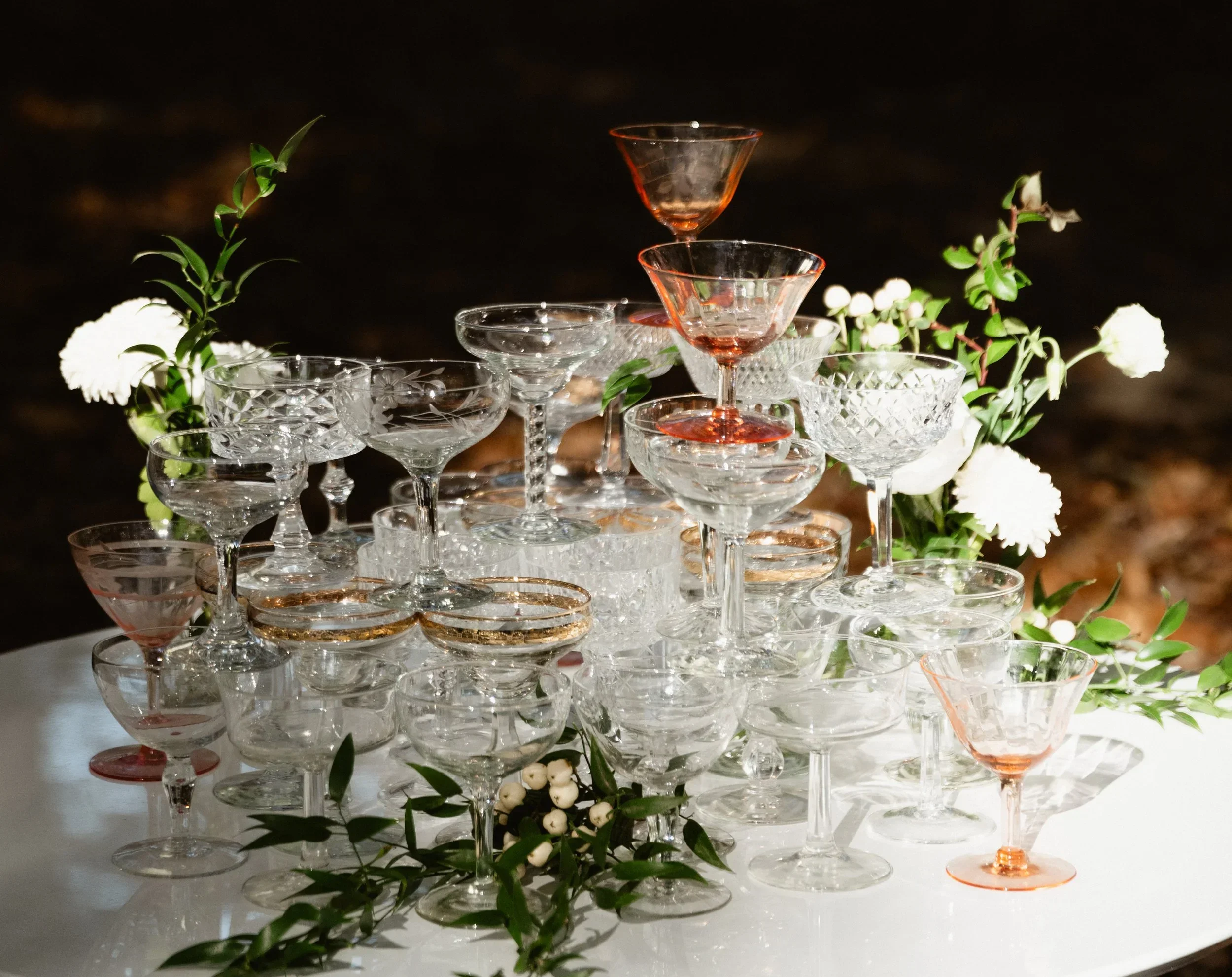 A pyramid of various crystal wine and cocktail glasses, decorated with white flowers and green foliage on a white table.