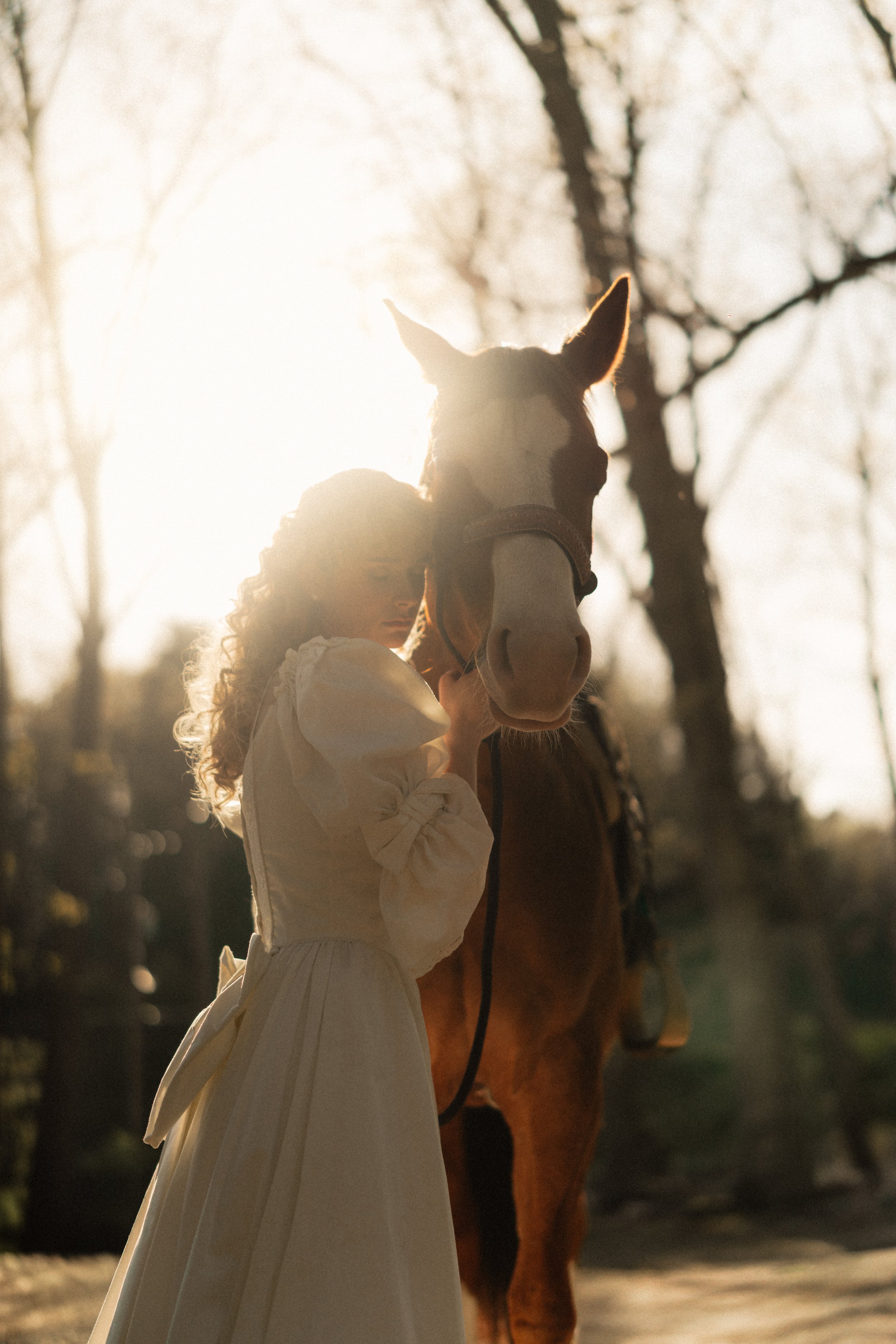 A woman in a white dress stands beside a horse with a white and brown face, the woman is leaning her head against the horse's neck during sunset or sunrise, with trees in the background.