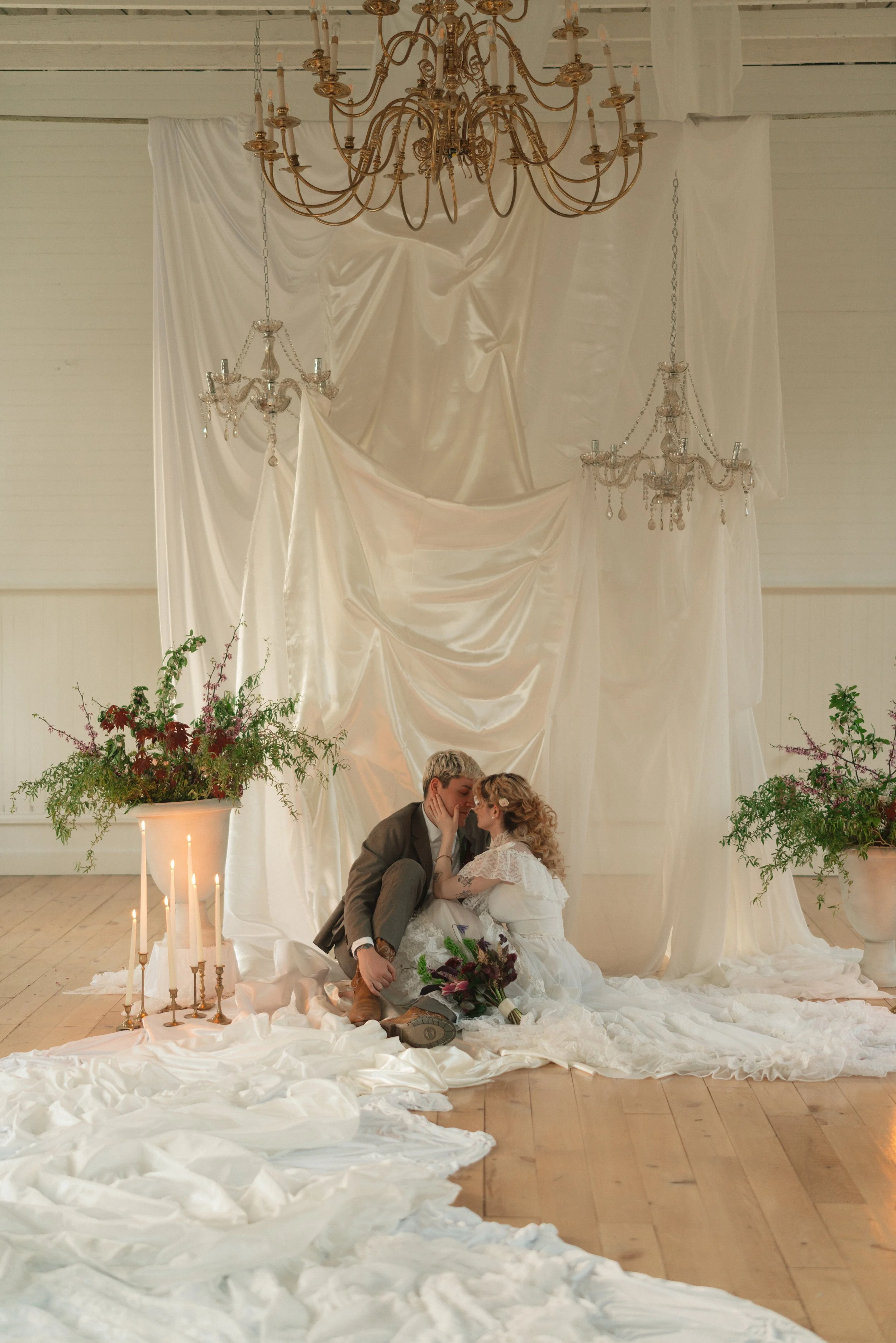 A couple in wedding attire sitting on the floor and touching foreheads, with a white draped backdrop, chandeliers, candles, and floral arrangements around them.