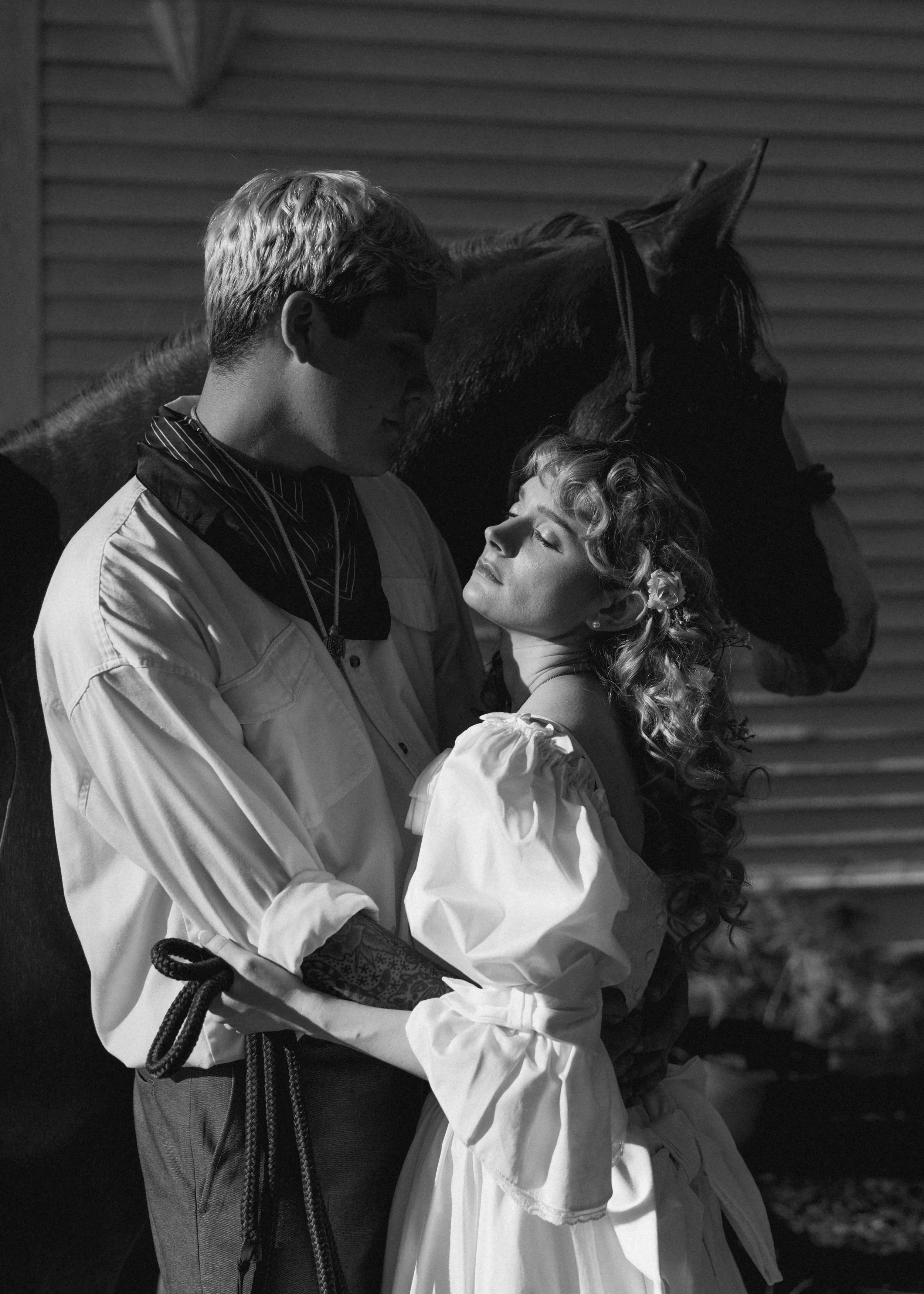 A black-and-white photo of a young man and woman in vintage clothing embracing with a horse nearby, with a barn in the background.