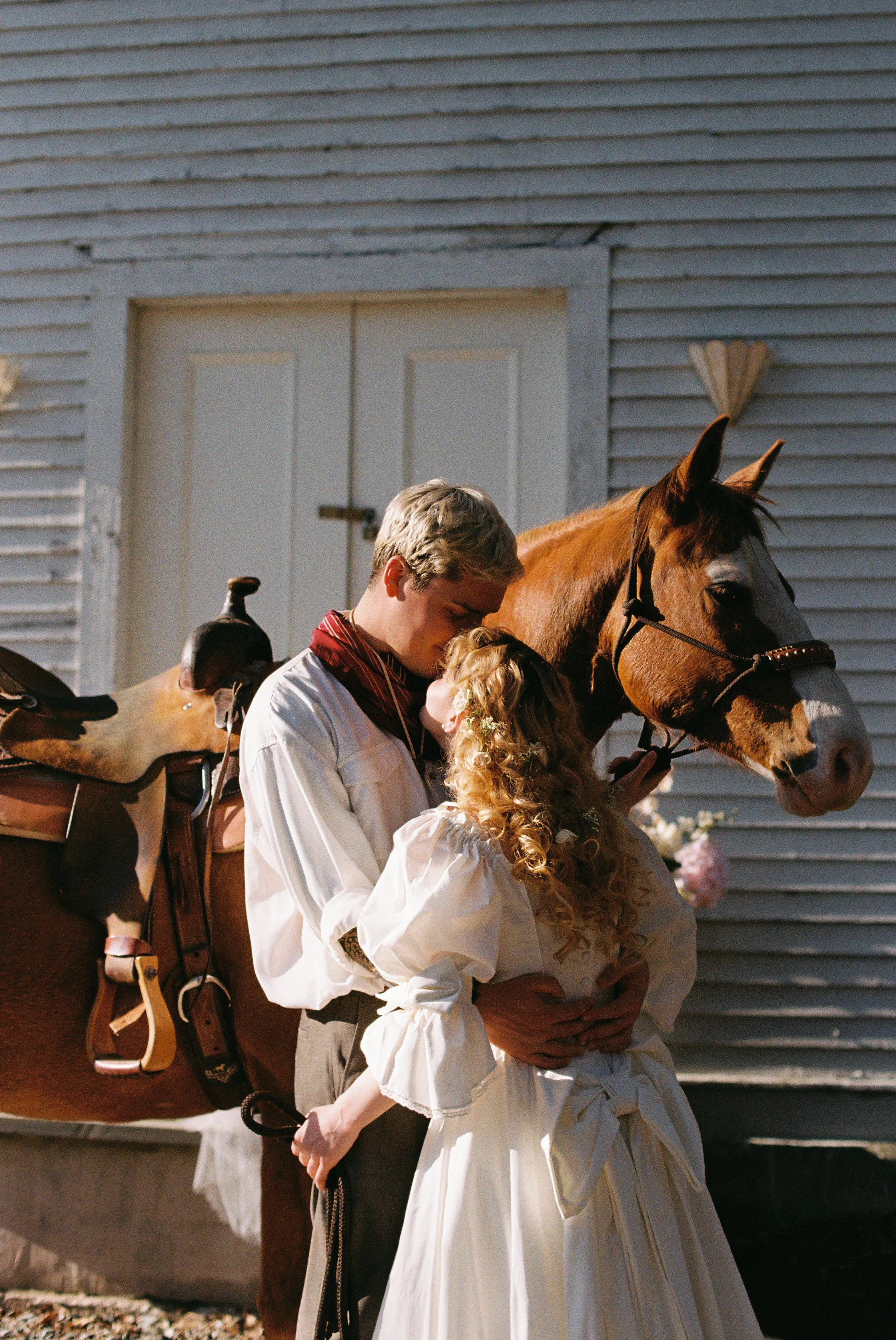 A man and a woman sharing a kiss, standing close to a horse outdoors, with a barn in the background.