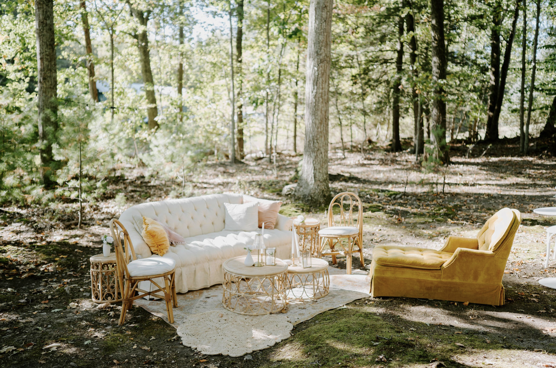 Outdoor seating arrangement in a wooded area with a white tufted sofa, a yellow velvet armchair, and wicker side tables, surrounded by trees and natural light.