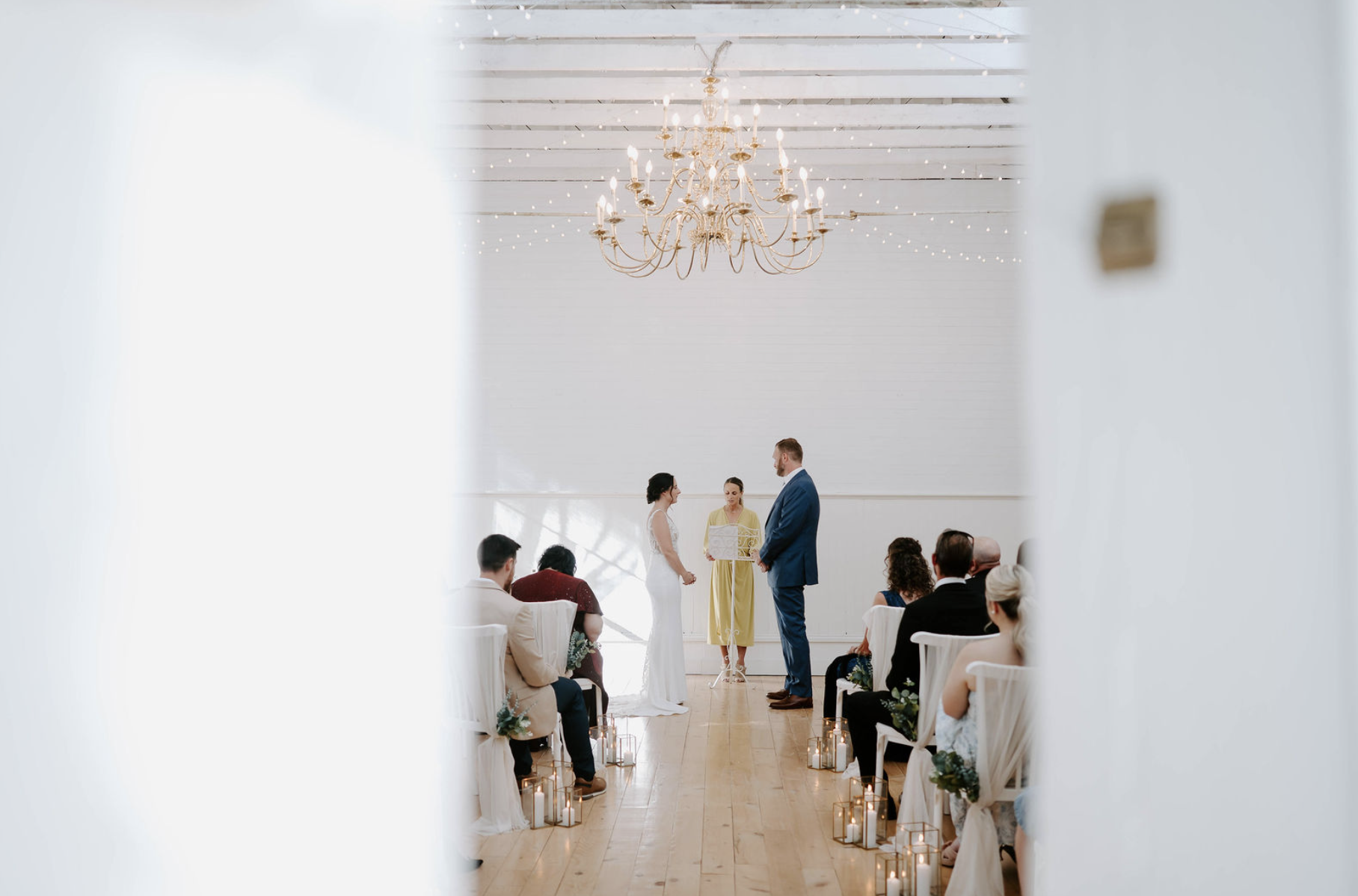 A wedding ceremony with the bride and groom holding hands before a officiant in a white chapel with a large chandelier and white wooden beams.