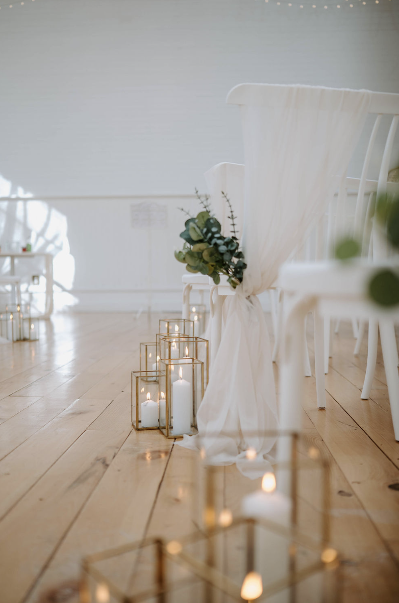 Elegant wedding ceremony setup with white chairs, white drapery, greenery, and candles in glass lanterns on a wooden floor.