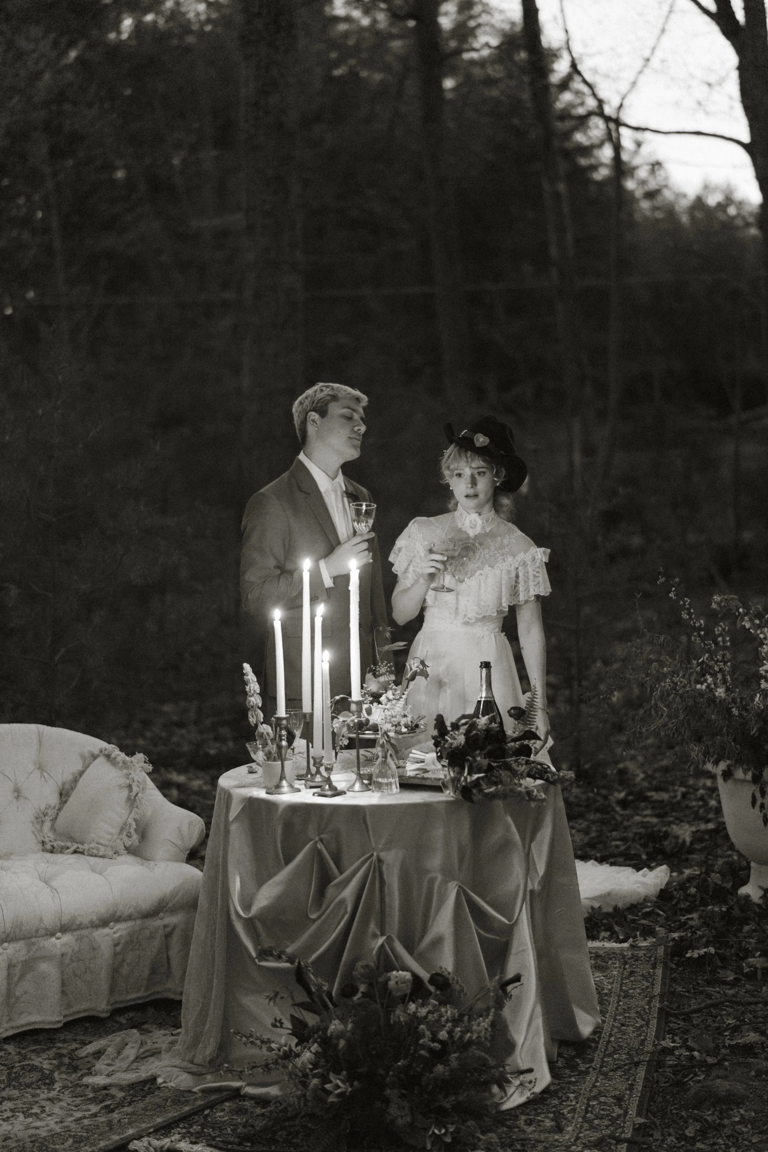 Two people, a man and a woman, dressed in vintage clothing, standing behind a decorated table with candles and flowers in an outdoor woodland setting at dusk.