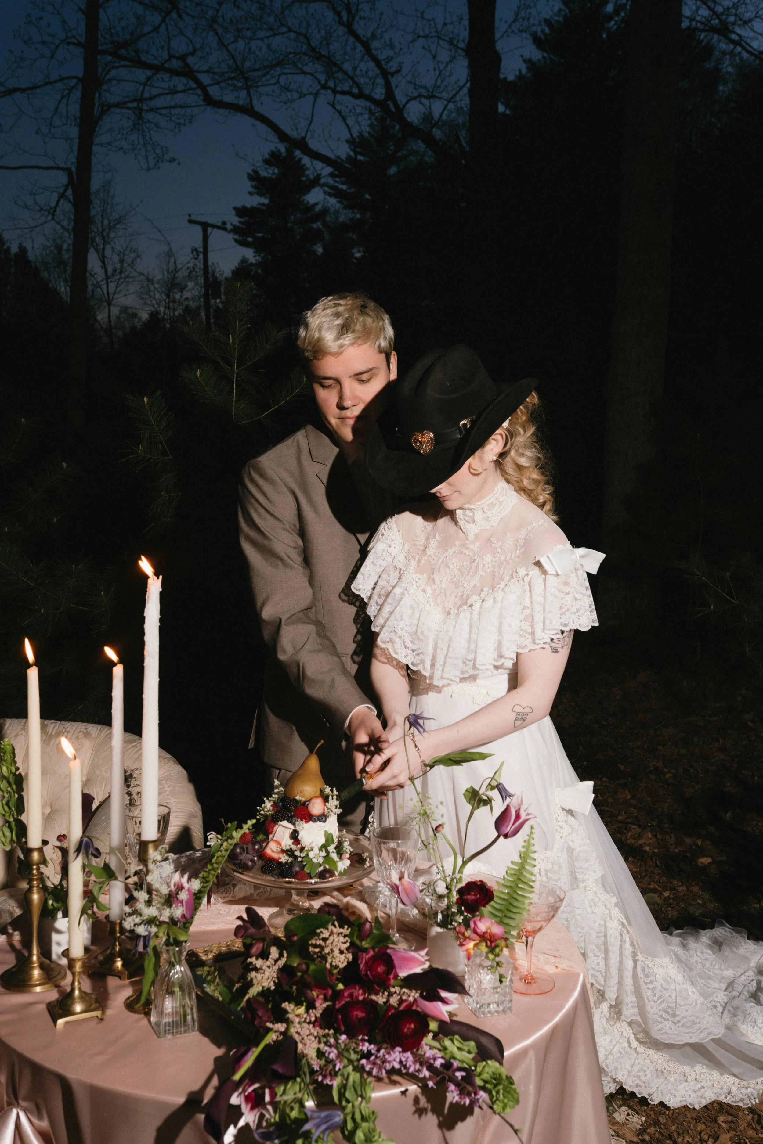 A couple is cutting a wedding cake outdoors at night, with candles and floral arrangements on the table.