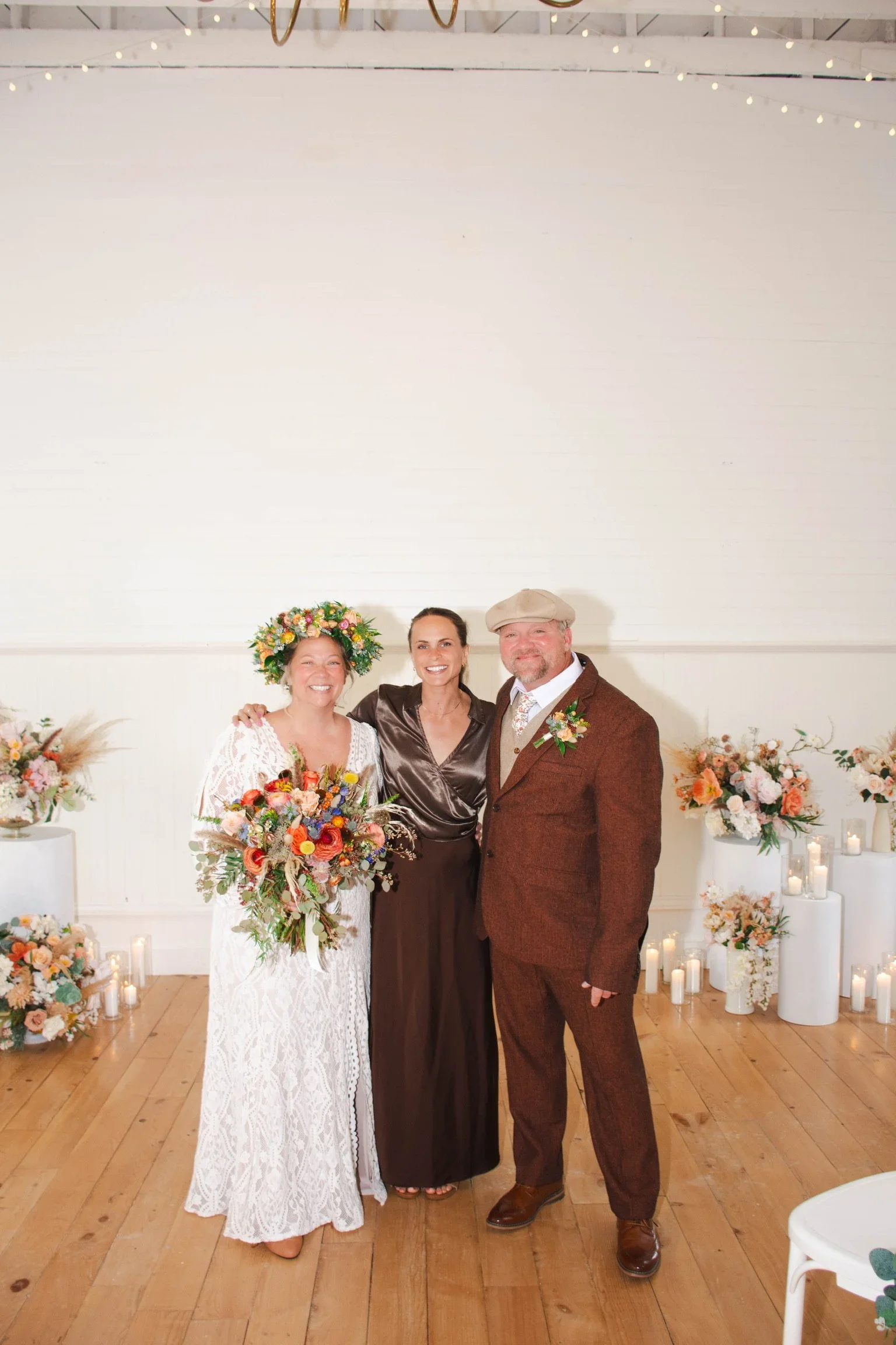 Three people standing together at a celebration, with decorated flowers and candles behind them. The woman on the left is holding a bouquet and wearing a floral crown, the woman in the middle is wearing a dark satin dress, and the man on the right is dressed in a brown suit with a flat cap.