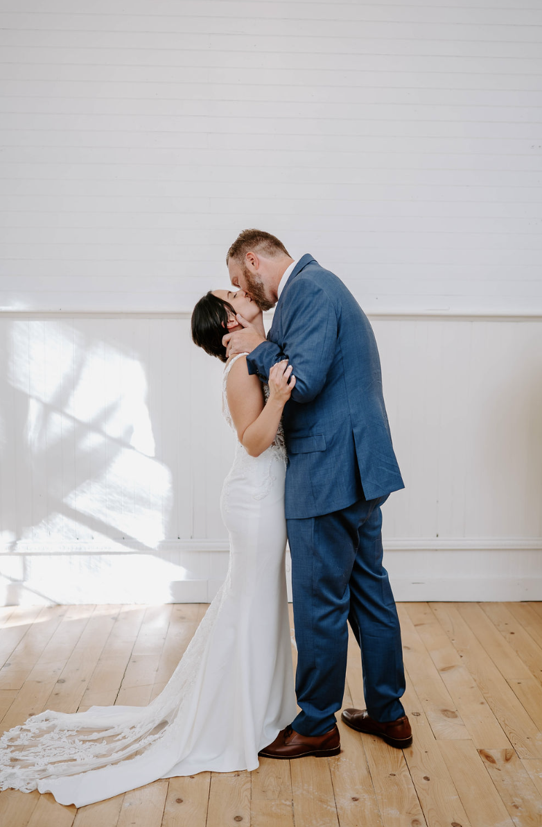 A couple sharing a kiss at their wedding, with the bride in a white gown and the groom in a blue suit, inside a bright room with white walls and wooden flooring.