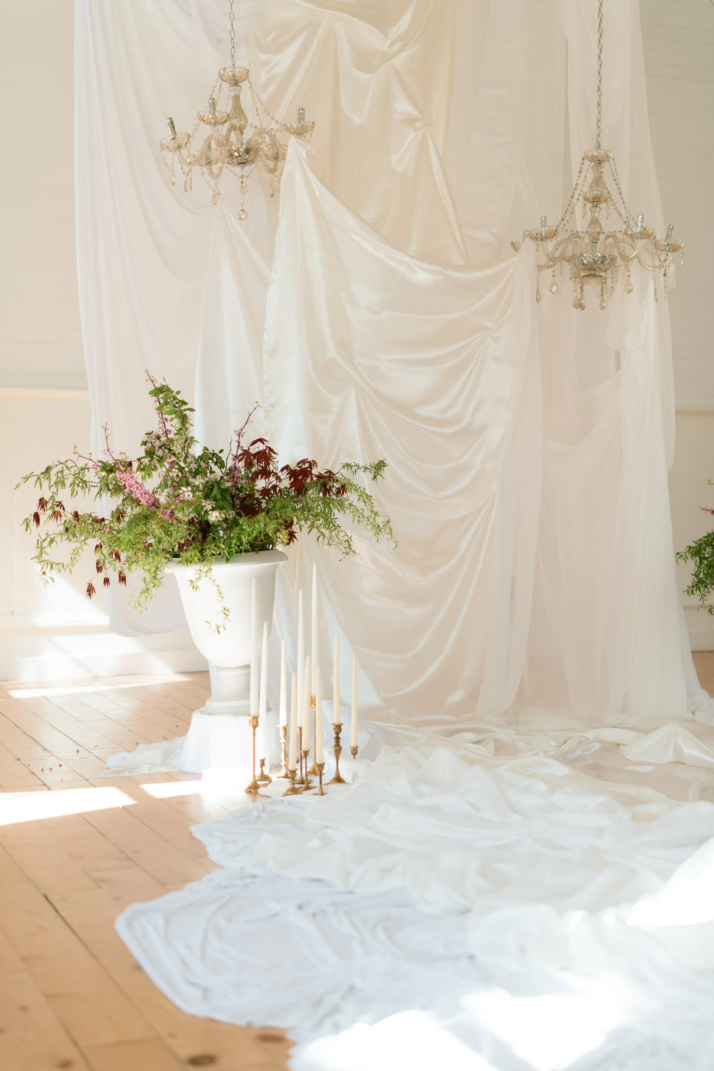 Elegant wedding or special event setup with white satin drapes, chandeliers, flowers in a white pot, and lit white candles on the floor.