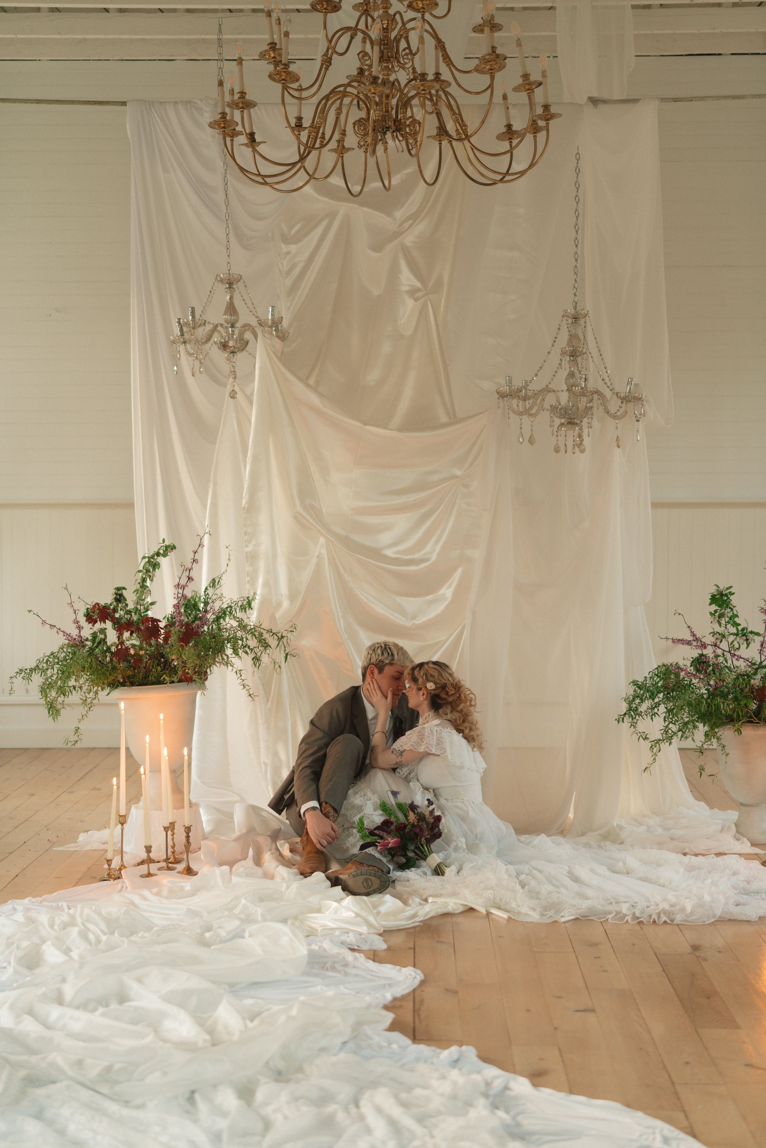 A couple dressed in wedding attire sitting on the floor close together, with their foreheads touching. They are surrounded by white fabric and candles, with large potted plants on either side. Chandeliers hang above them, and the background features white draped fabric.