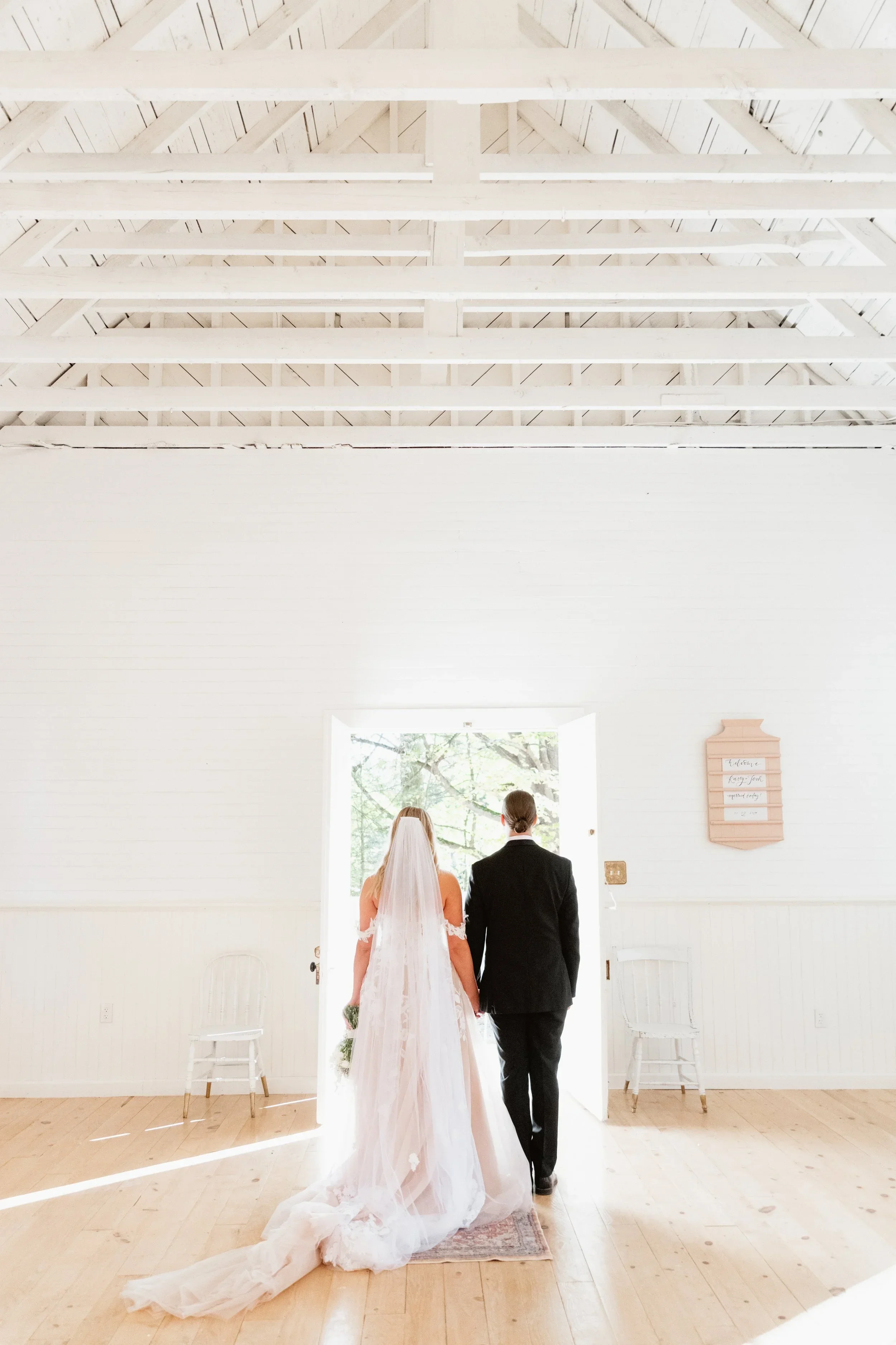 A bride in a wedding dress and veil and a groom in a black suit walking out of a bright, white room through a doorway to an outdoor setting with trees.