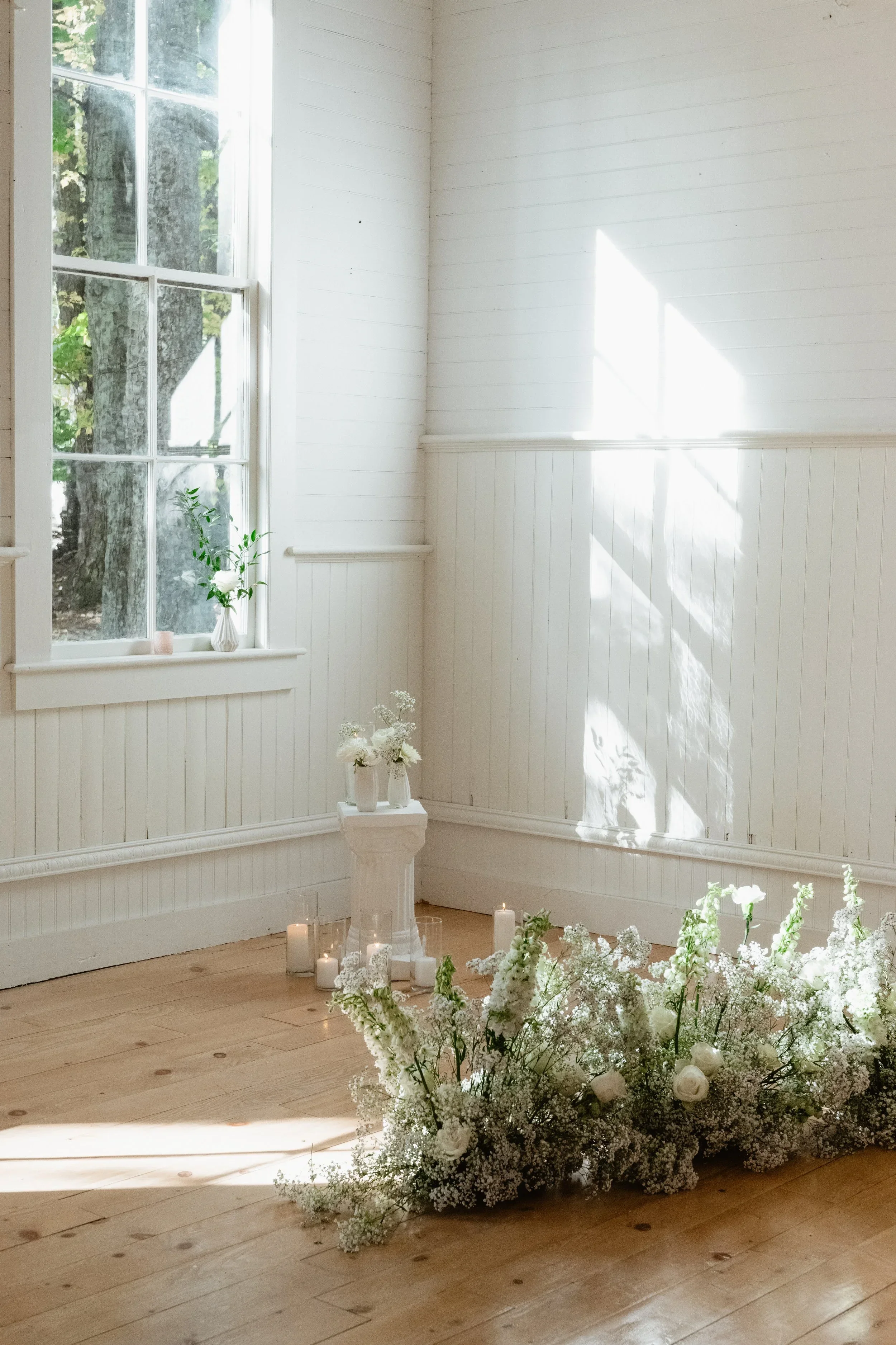 A bright, minimalistic room with white wooden walls and a window allowing natural light. The floor is wooden, and the room is decorated with white flowers and candles, suggesting a setting for a wedding or special event.