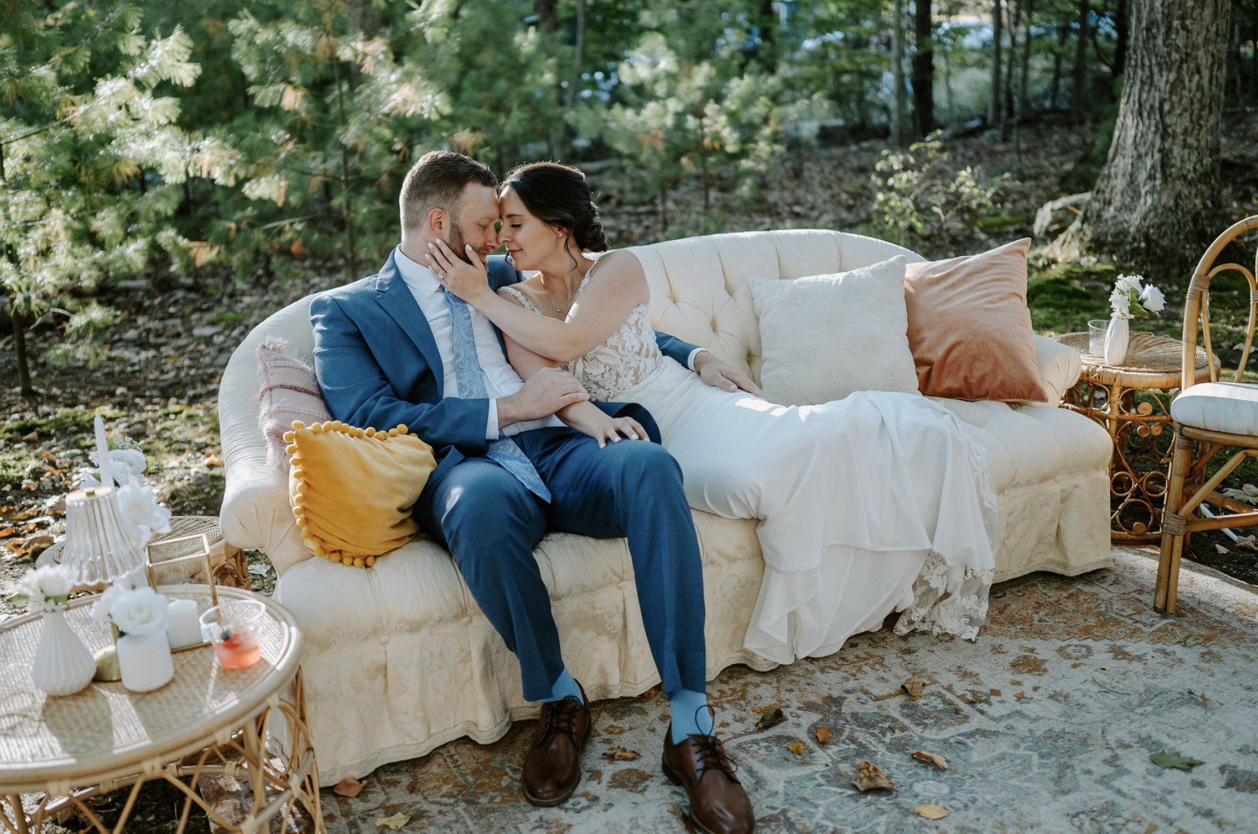 A bride and groom sitting close together on a vintage sofa outdoors surrounded by trees, sharing a tender moment with eyes closed and faces touching