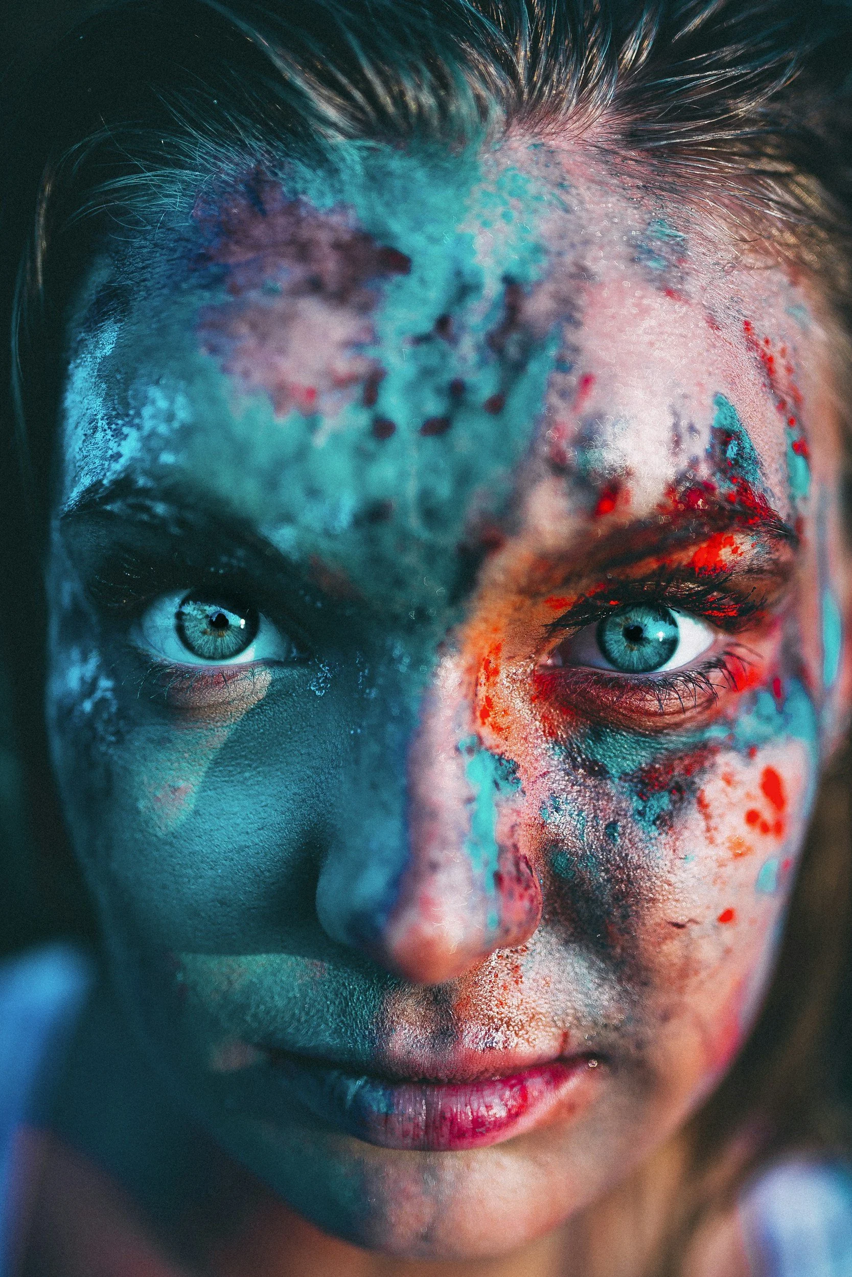 Close-up of a woman's face with paint splattered in blue, red, and white, emphasizing her blue eyes and intense gaze.