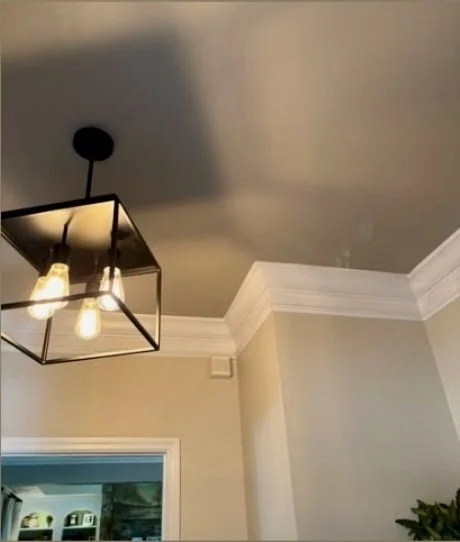 Close-up of a ceiling with a modern black geometric light fixture and two exposed light bulbs, and part of an interior wall with crown molding.
