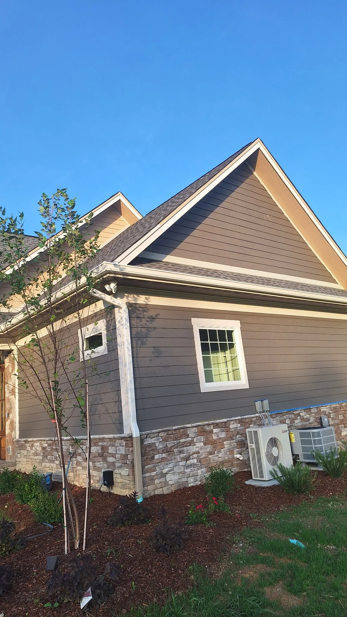 Exterior view of a house with gray siding, stone foundation, small window, and HVAC units outside, with a clear blue sky and some landscaping.
