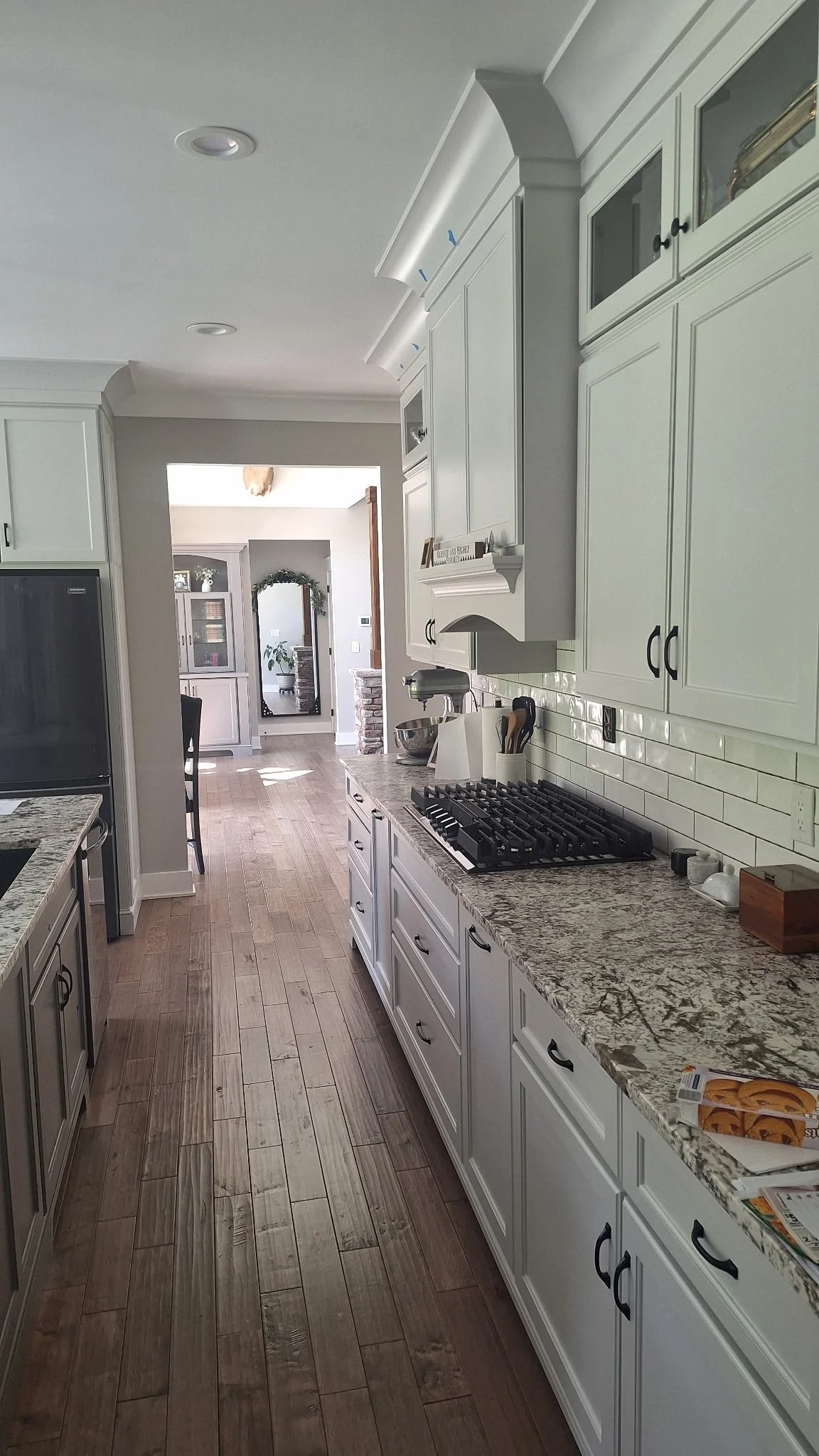 A bright, modern kitchen with white cabinetry and granite countertops. The image shows a view from the side, with a focus on the countertop, stove, and upper cabinets. There is a doorway leading to a dining area and living room in the background.