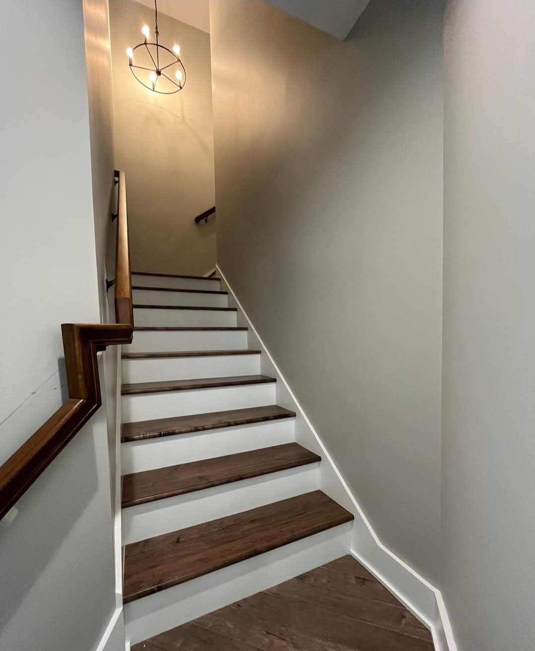 A staircase with wooden steps and a wooden handrail, leading to a second floor, illuminated by a wall-mounted circular light fixture.
